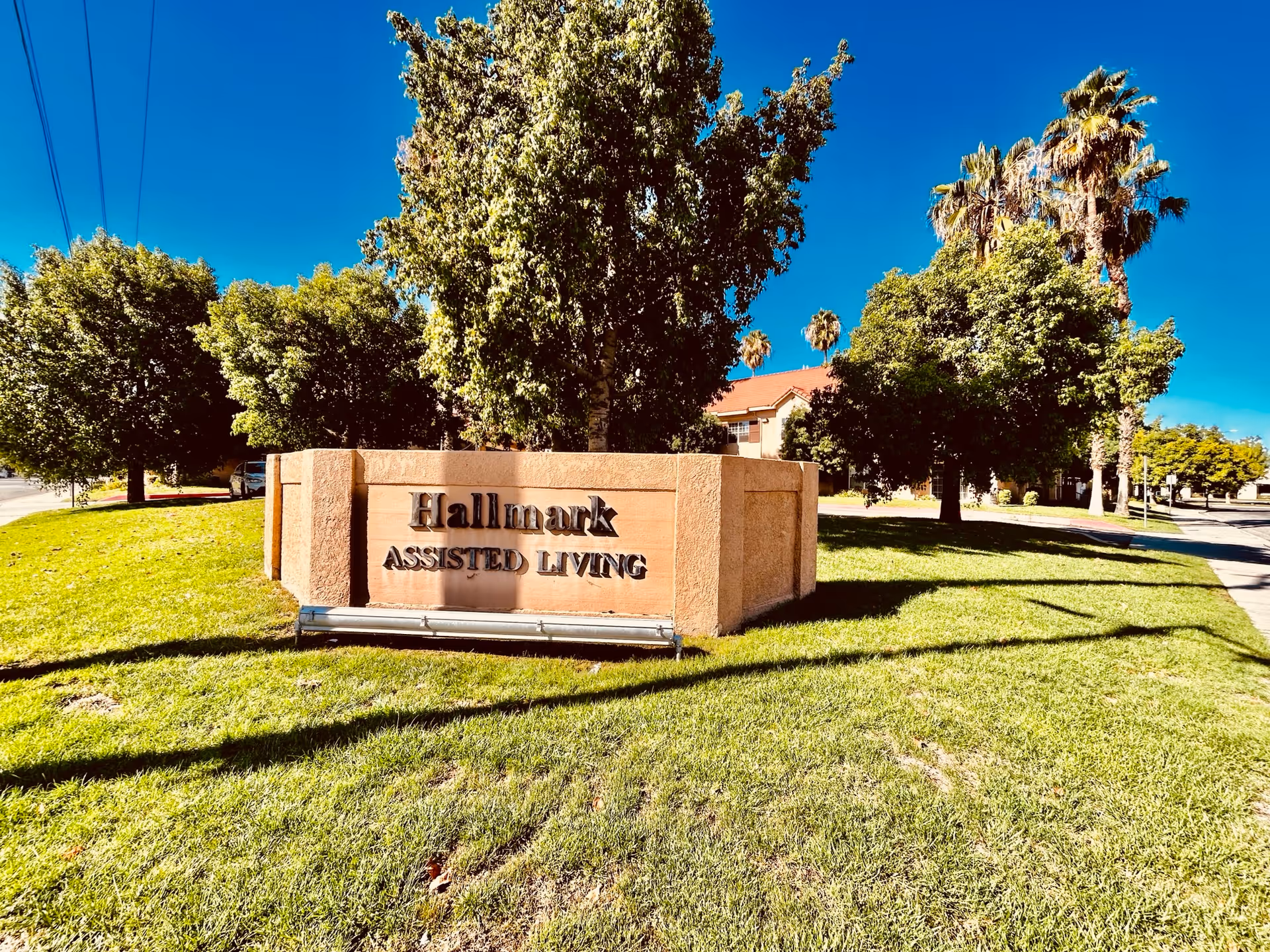 Outdoor view of a sign for Hallmark Assisted Living on a grassy area with trees and a clear blue sky in the background.
