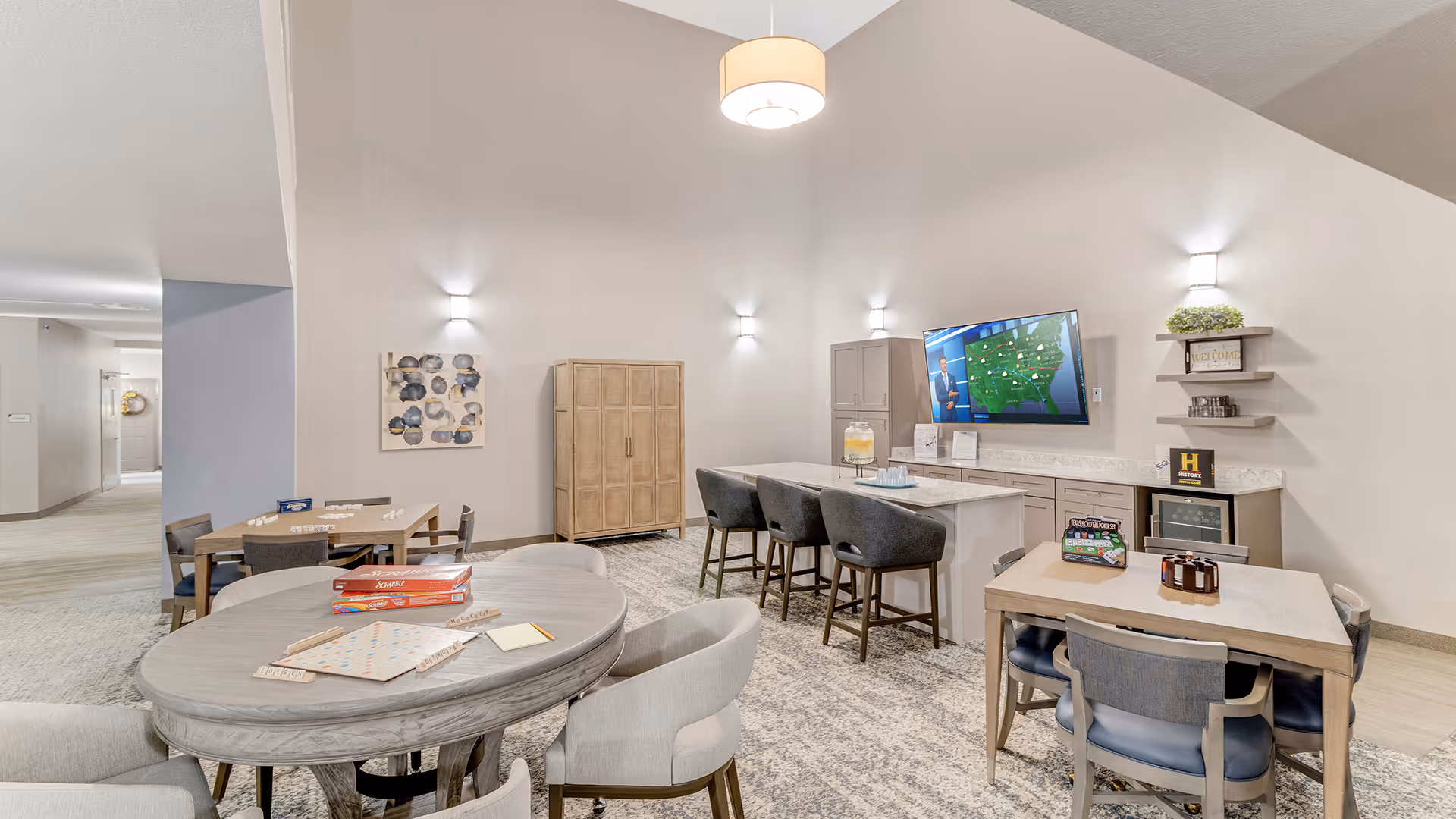 A bright and spacious common area in a senior living facility featuring multiple tables and chairs arranged for social activities. There is a large flat-screen TV mounted on the wall displaying a weather forecast, a countertop with stools, and shelves with decorative items. The room has soft lighting and neutral-colored walls and flooring.