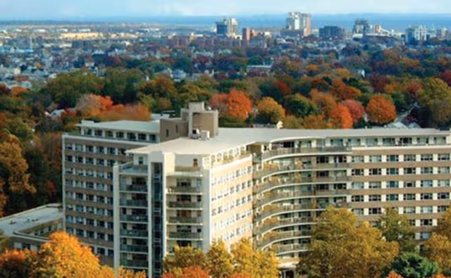 A large, multi-story senior living building surrounded by autumn-colored trees.