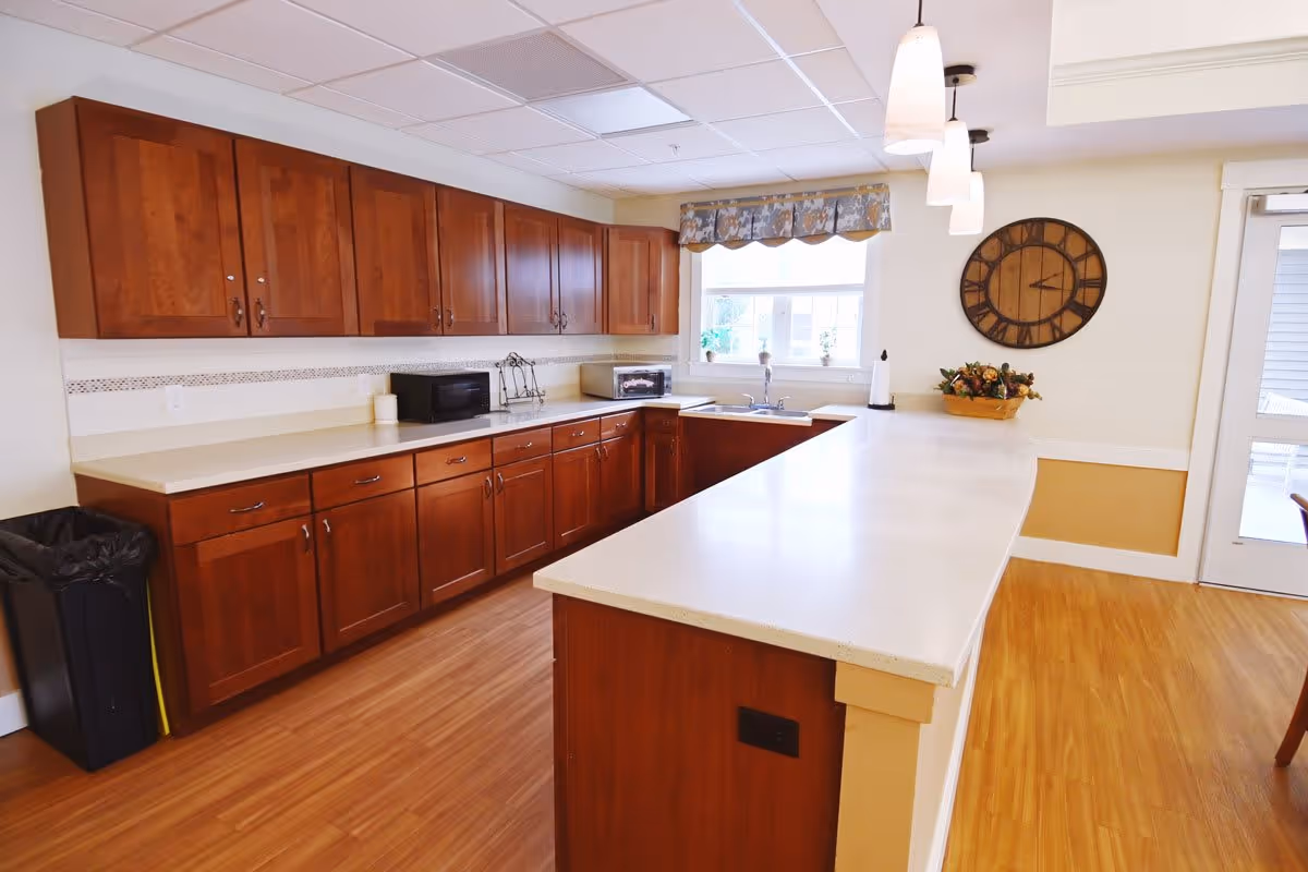 A bright kitchen area with wooden cabinets, a long white countertop island, a sink under a window with a floral valance, a wall clock, and three pendant lights hanging from the ceiling. The floor is wooden, and there is a trash bin on the left side.
