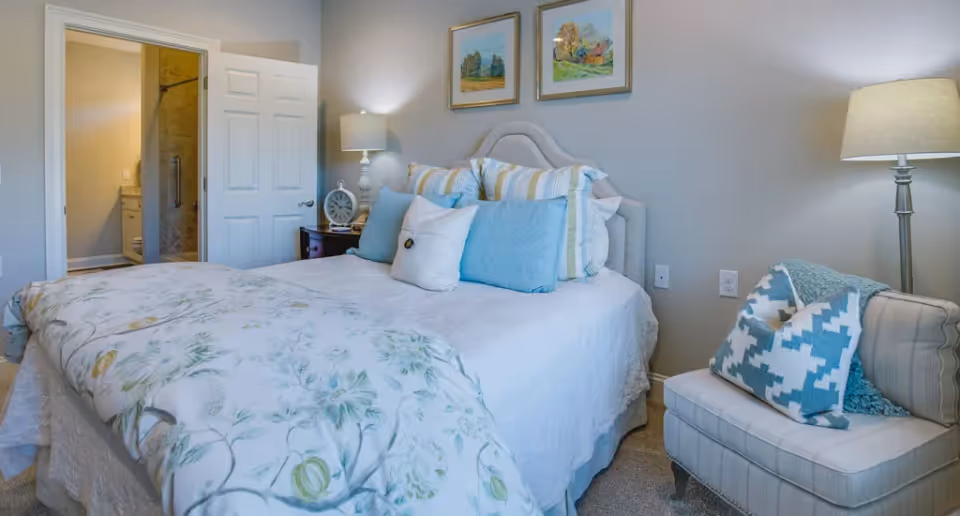 A neatly made bed with white and floral bedding in a bedroom. The bed has multiple pillows in shades of blue and white. To the right of the bed is a cushioned chair with a blue and white patterned pillow and a blue throw blanket. A floor lamp stands next to the chair. On the left side of the bed, there is a nightstand with a lamp and a clock. The bedroom door is open, revealing a bathroom with a shower and vanity. Two framed paintings hang on the wall above the bed.