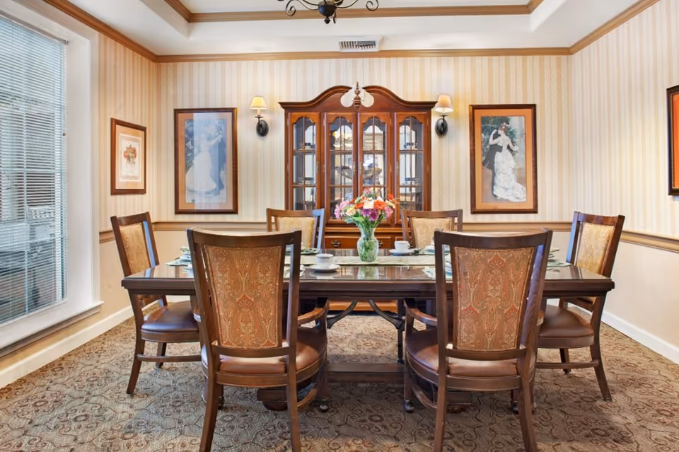 Dining room with a wooden table set for meals, upholstered chairs, a china cabinet, framed artwork, and a vase of flowers.
