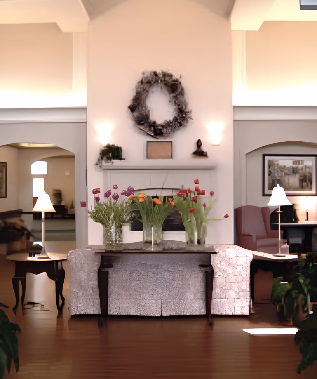 A cozy living room area with a stone fireplace in the center, decorated with a wreath above the mantel and three vases of colorful flowers on the mantel. There are two side tables with lamps on either side of the fireplace, and armchairs and framed artwork are visible in the background. The room has warm lighting and wooden flooring.