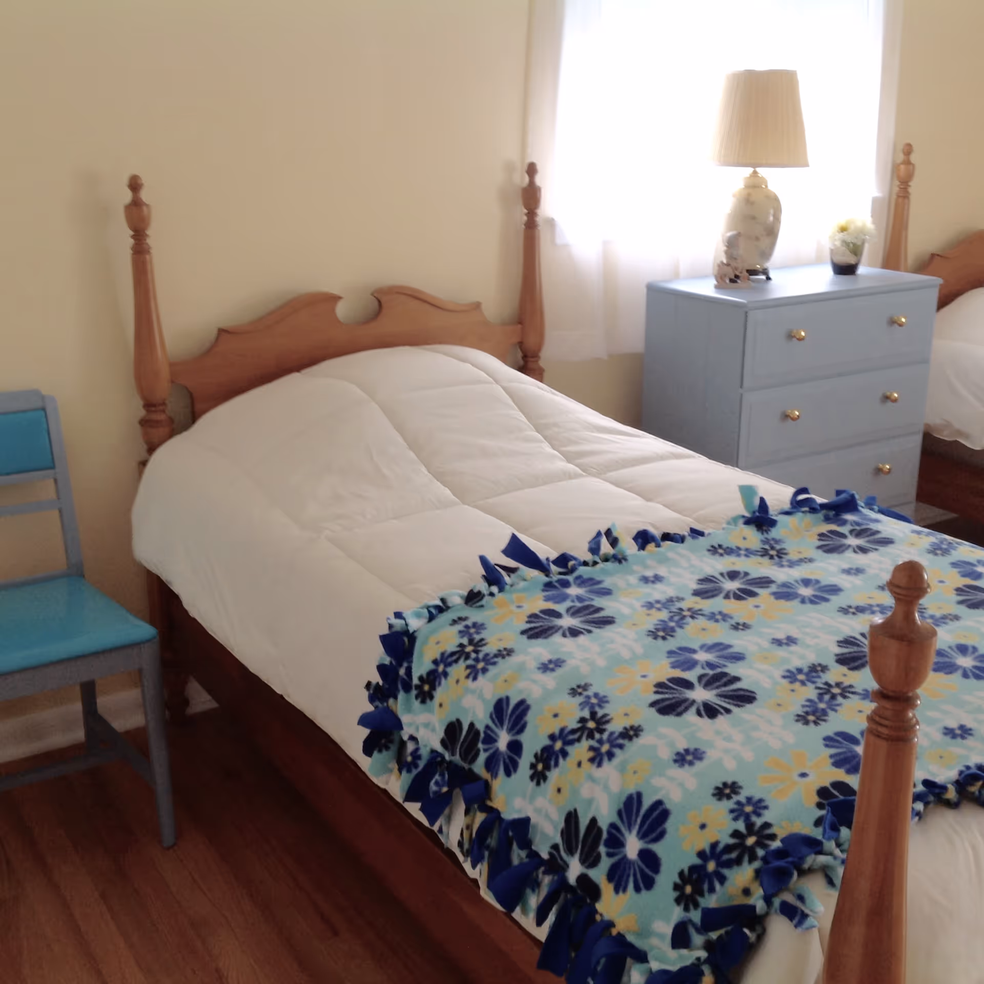 A bedroom with a wooden bed frame, a white comforter, and a blue floral blanket at the foot of the bed. Next to the bed is a blue chair and a light blue dresser with a lamp and a small plant on top. Light filters in through a window with white curtains.