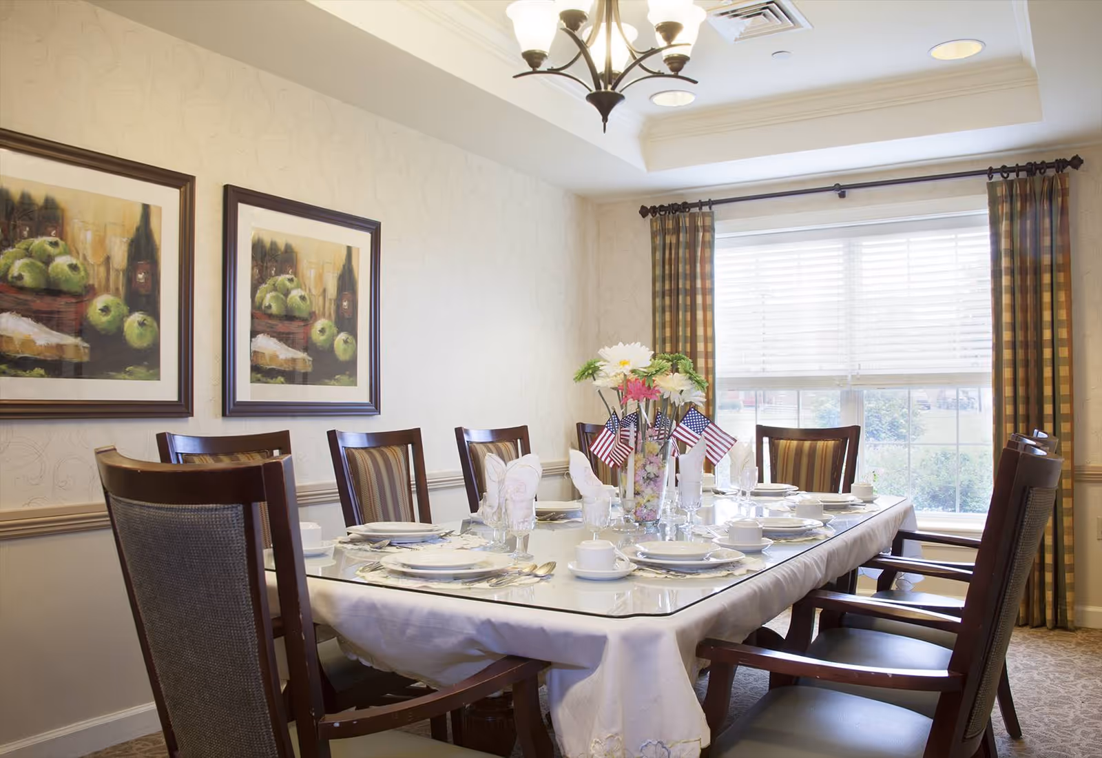 A formal dining room with a long rectangular table set for a meal. The table is covered with a white tablecloth and has plates, cups, glasses, and silverware arranged neatly. A vase with flowers and small American flags is placed in the center of the table. The room has patterned curtains on the window, two framed paintings of green apples on the wall, and a chandelier hanging from the ceiling.