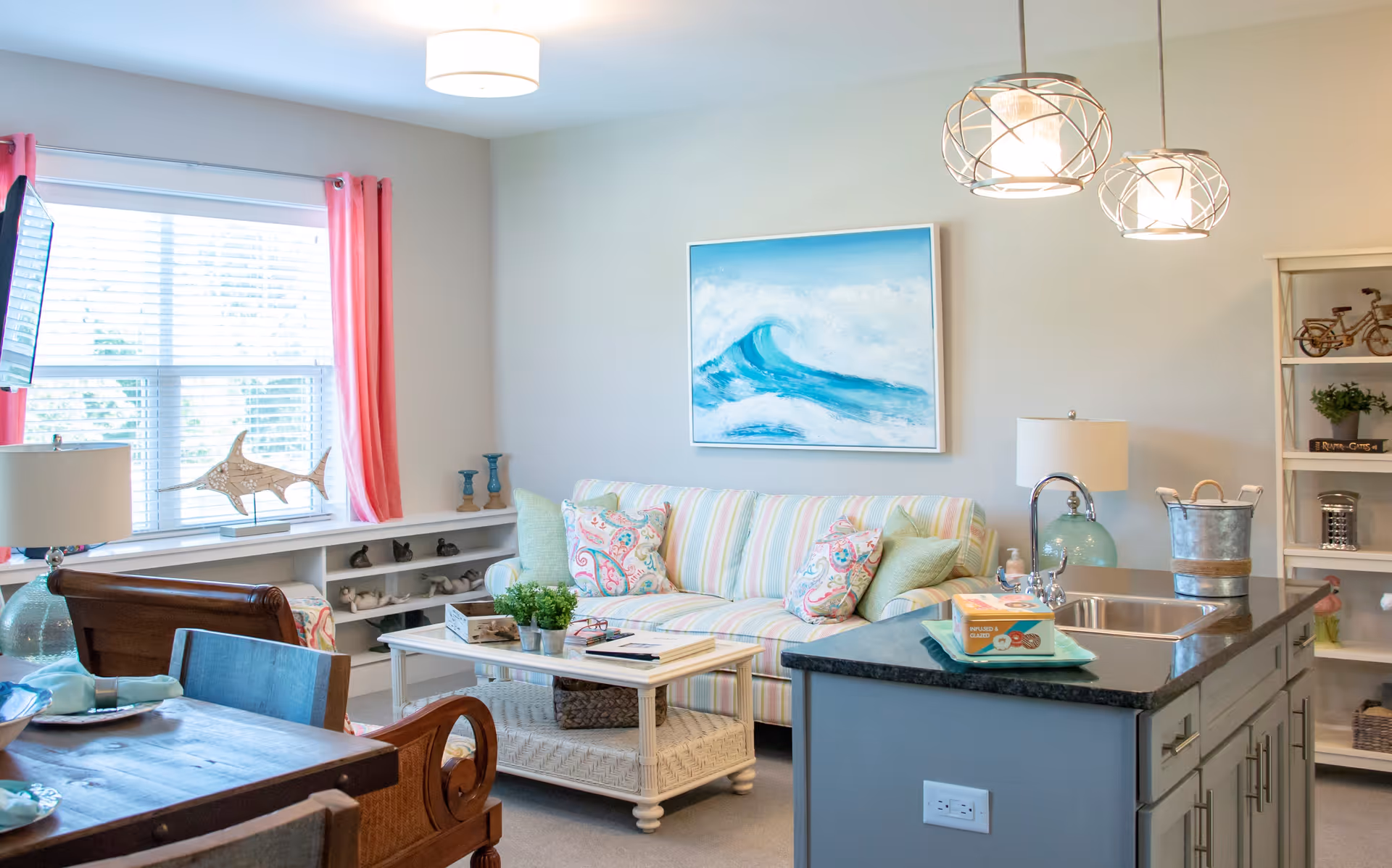 Bright and cozy living room with a striped couch adorned with colorful pillows, a white coffee table with decorative items, and a kitchen island with a sink and pendant lights above. A large window with pink curtains lets in natural light, and a painting of an ocean wave hangs on the wall.
