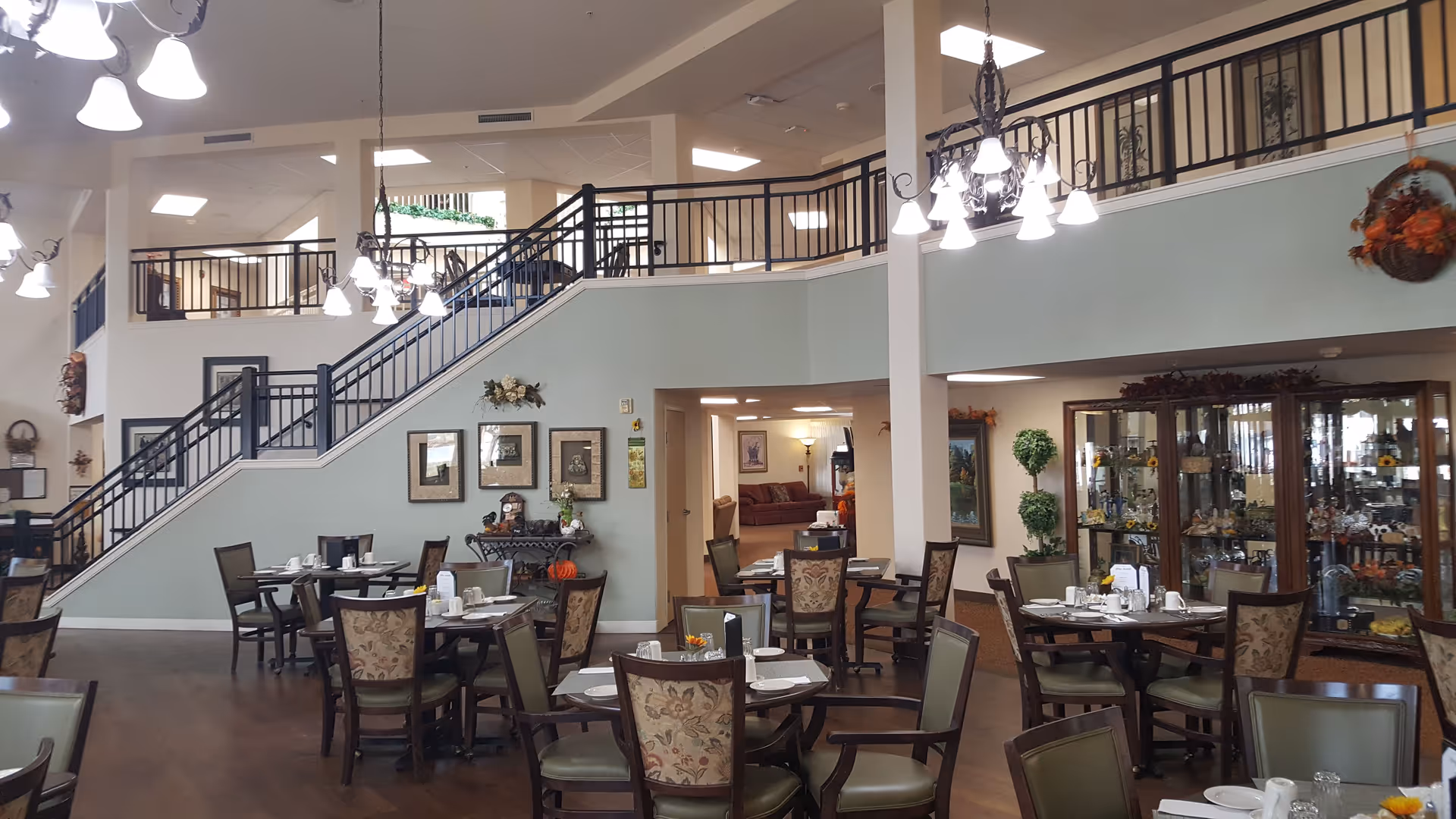 Spacious senior living dining room with multiple set tables, chandeliers, and a central staircase leading to an upper balcony.