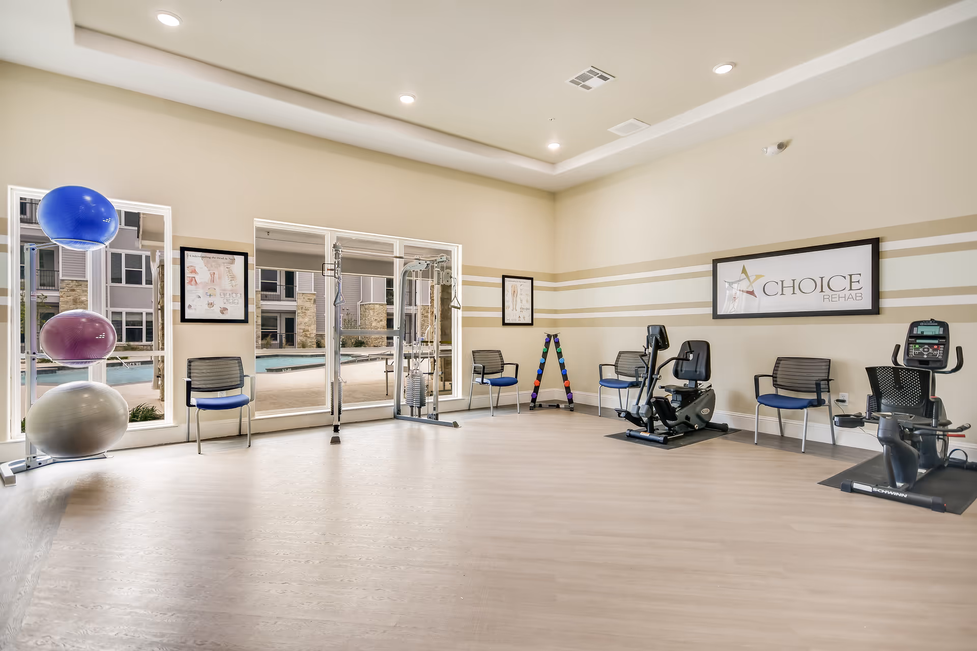 A bright and spacious rehabilitation exercise room with large windows showing an outdoor pool area. The room contains exercise equipment including stationary bikes, a multi-functional weight machine, colorful exercise balls stacked on a rack, and a set of dumbbells on a rack. Several chairs are placed along the walls, and a framed sign on the wall reads 'CHOICE REHAB'.