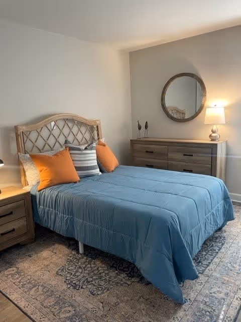 A neatly made bed with a blue comforter and four pillows, two orange and two striped, in a bedroom. The bed has a wooden headboard with a lattice design. Next to the bed is a wooden nightstand with a lamp. Across from the bed is a wooden dresser with a round mirror above it and a lamp on the right side. The floor has a patterned rug.