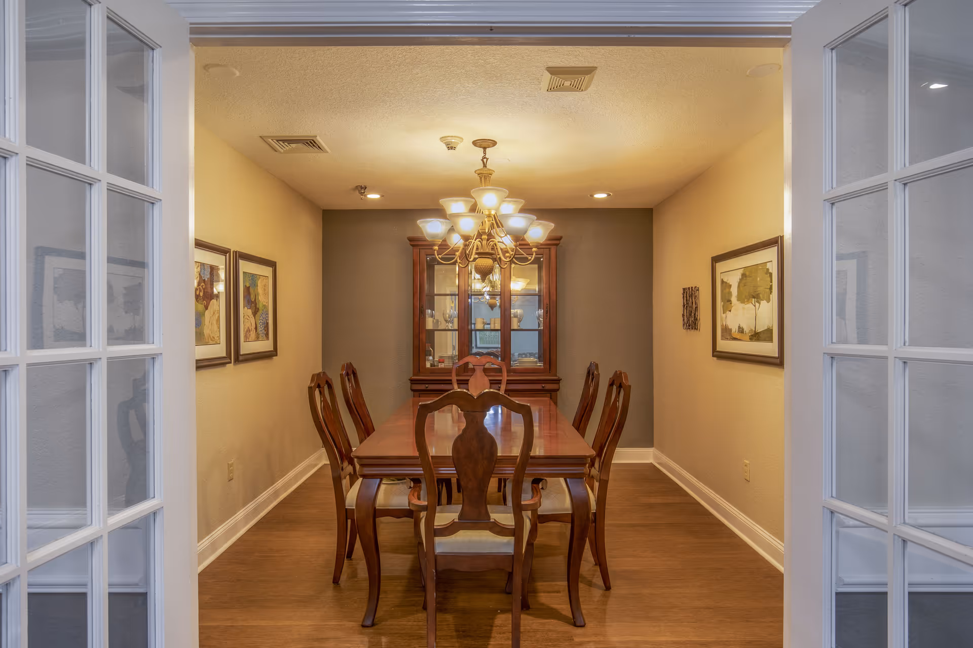 View through glass-paneled double doors into a dining room with a wooden dining table and six matching chairs. A chandelier with multiple lights hangs above the table. On the back wall, there is a wooden china cabinet with glass doors displaying dishes. The walls are decorated with framed artwork, and the floor is wooden.