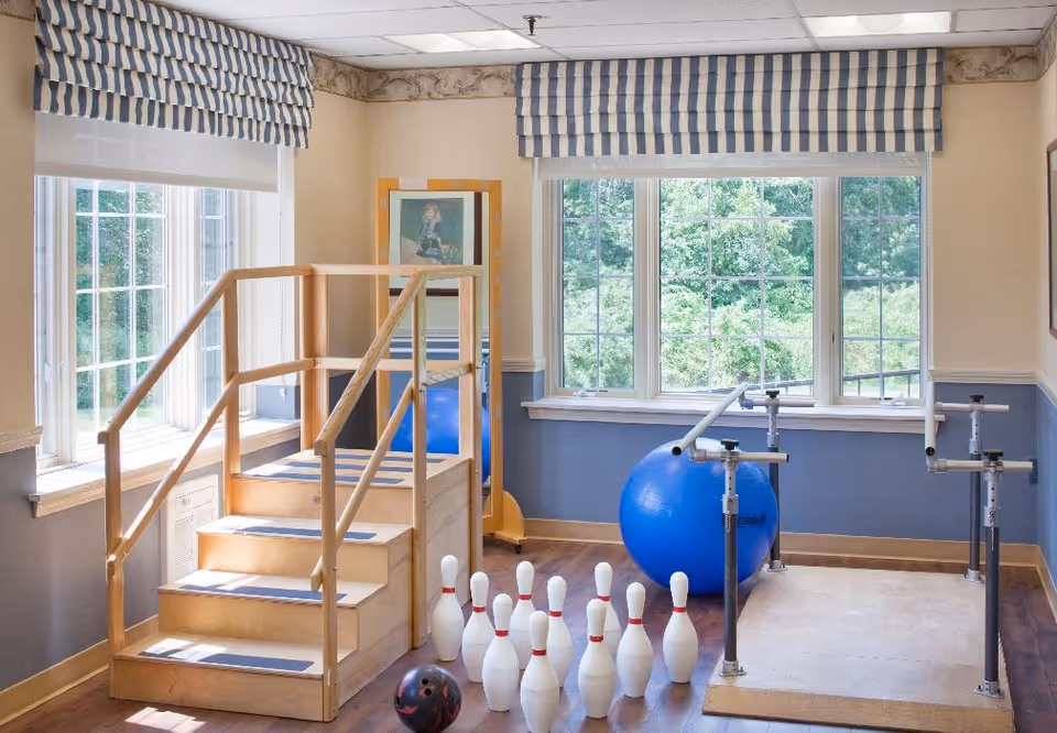 A bright therapy room with large windows showing greenery outside. The room contains wooden stairs with handrails for physical therapy, a large blue exercise ball, parallel bars for walking support, and a set of bowling pins with a bowling ball on the floor.