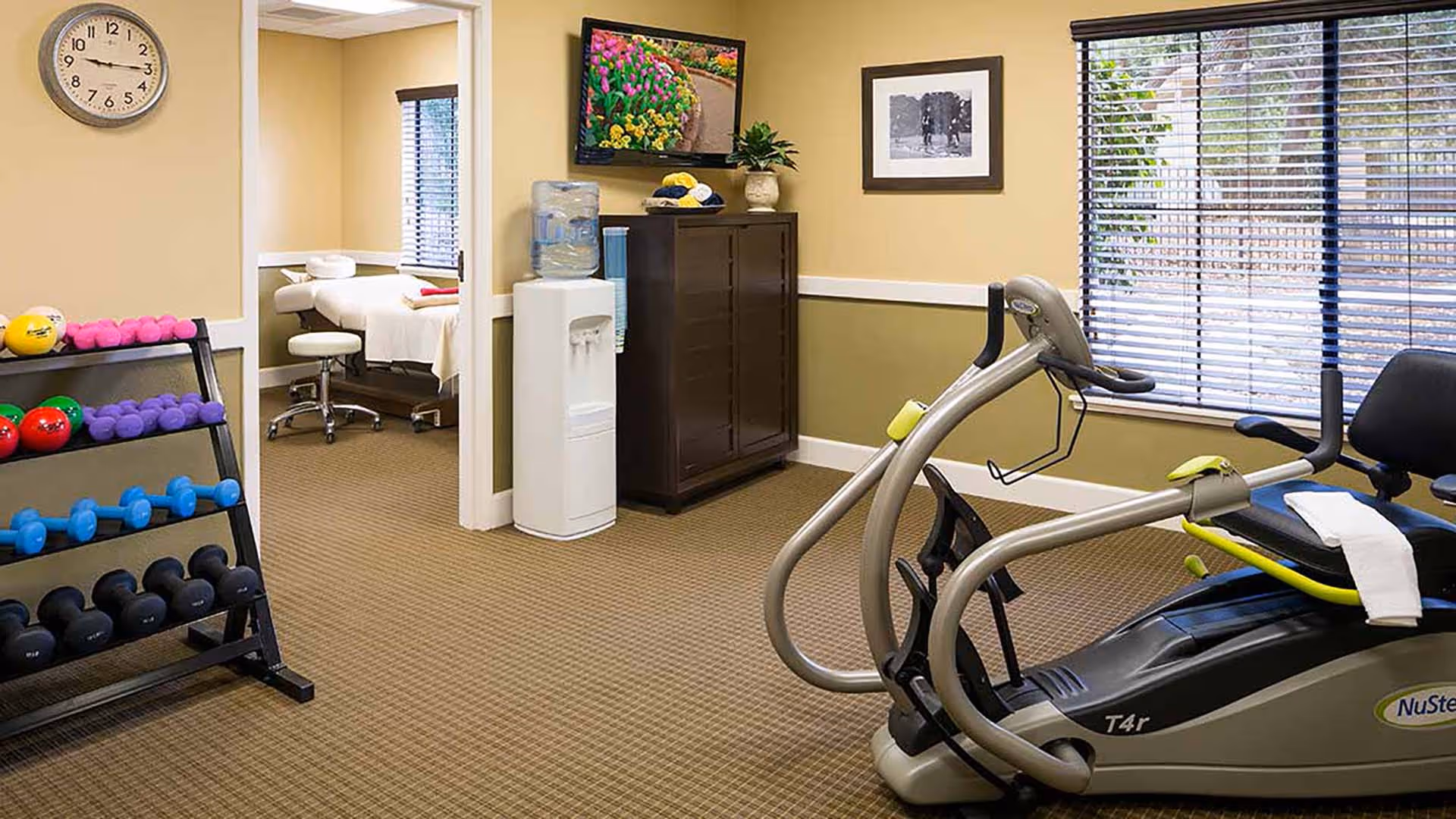 Fitness room with a NuStep exercise machine, a rack of dumbbells, a water cooler, and a treatment table visible through an open doorway.