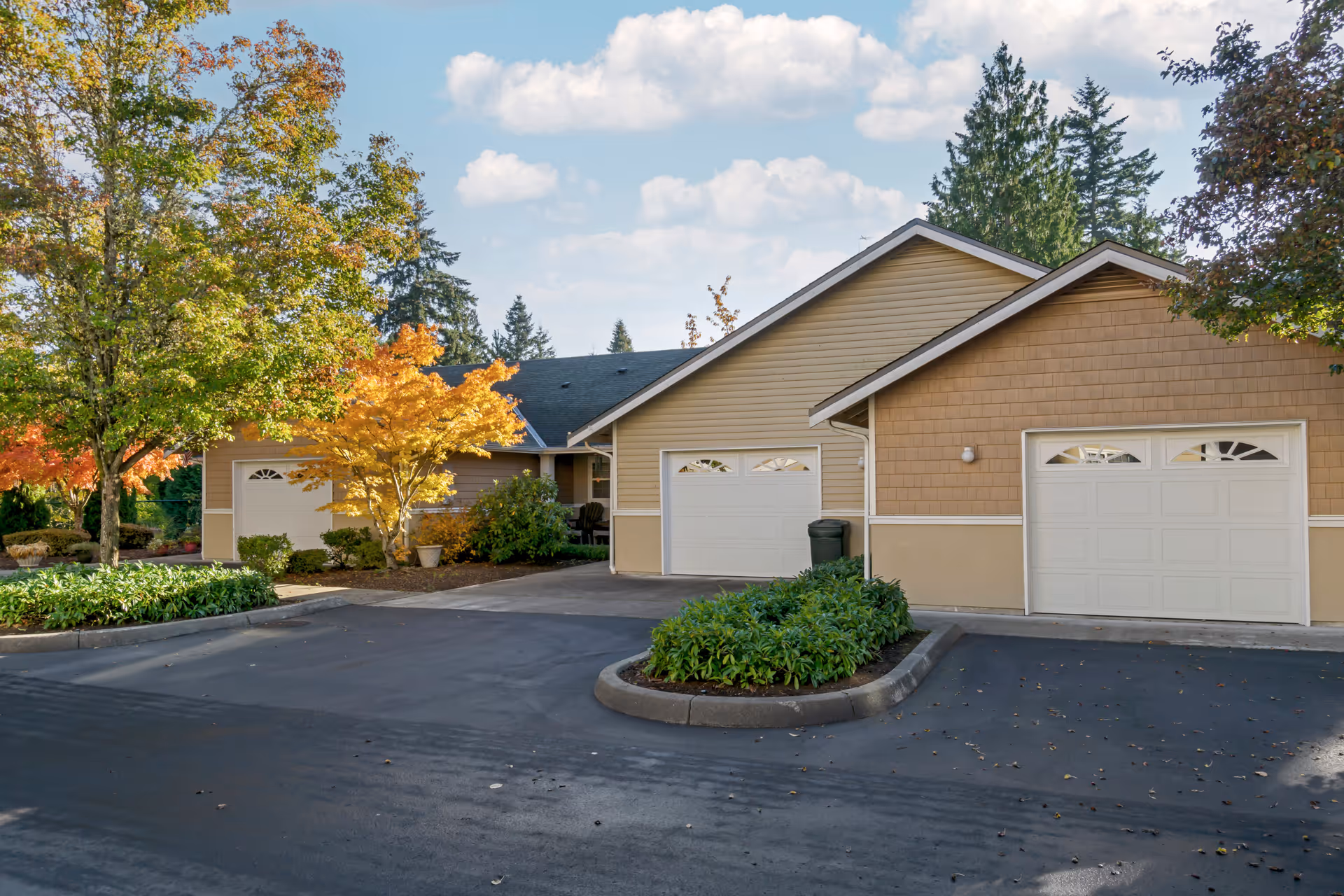 Exterior view of a residential building with beige siding and white garage doors, surrounded by trees with autumn foliage and landscaped greenery under a partly cloudy sky.