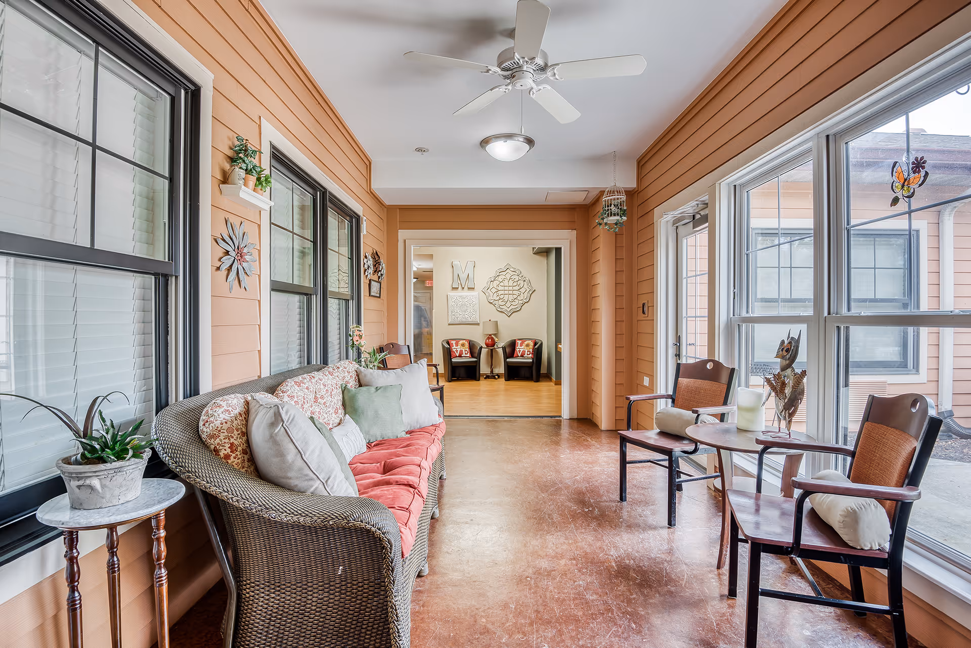 A bright indoor sitting area with large windows on the right side and peach-colored walls. There is a wicker sofa with floral and solid-colored cushions on the left, a small round side table with a potted plant, and two wooden chairs with cushions and a small table between them on the right. A ceiling fan and light fixture are mounted on the white ceiling. In the background, an adjoining room has two black chairs with red pillows spelling 'LOVE' and wall decorations including a large letter 'M'.