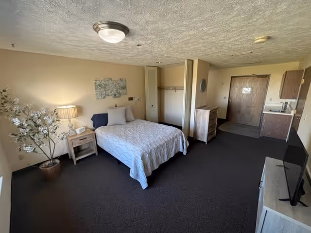A spacious bedroom in a retirement living facility featuring a neatly made bed with white bedding, a wooden nightstand with a lamp and clock, a potted plant with white flowers, an open closet, a wooden dresser, a small kitchenette area with a sink, and a television on a stand. The room has beige walls and dark carpet flooring.