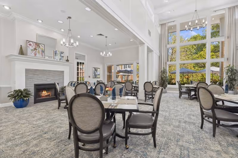 A bright and spacious dining room with multiple round tables and chairs arranged neatly. The room features a large window wall allowing natural light to flood in, showing a view of outdoor umbrellas and greenery. There is a fireplace on the left side with a decorative mantle, plants in blue pots, and elegant chandeliers hanging from the ceiling.