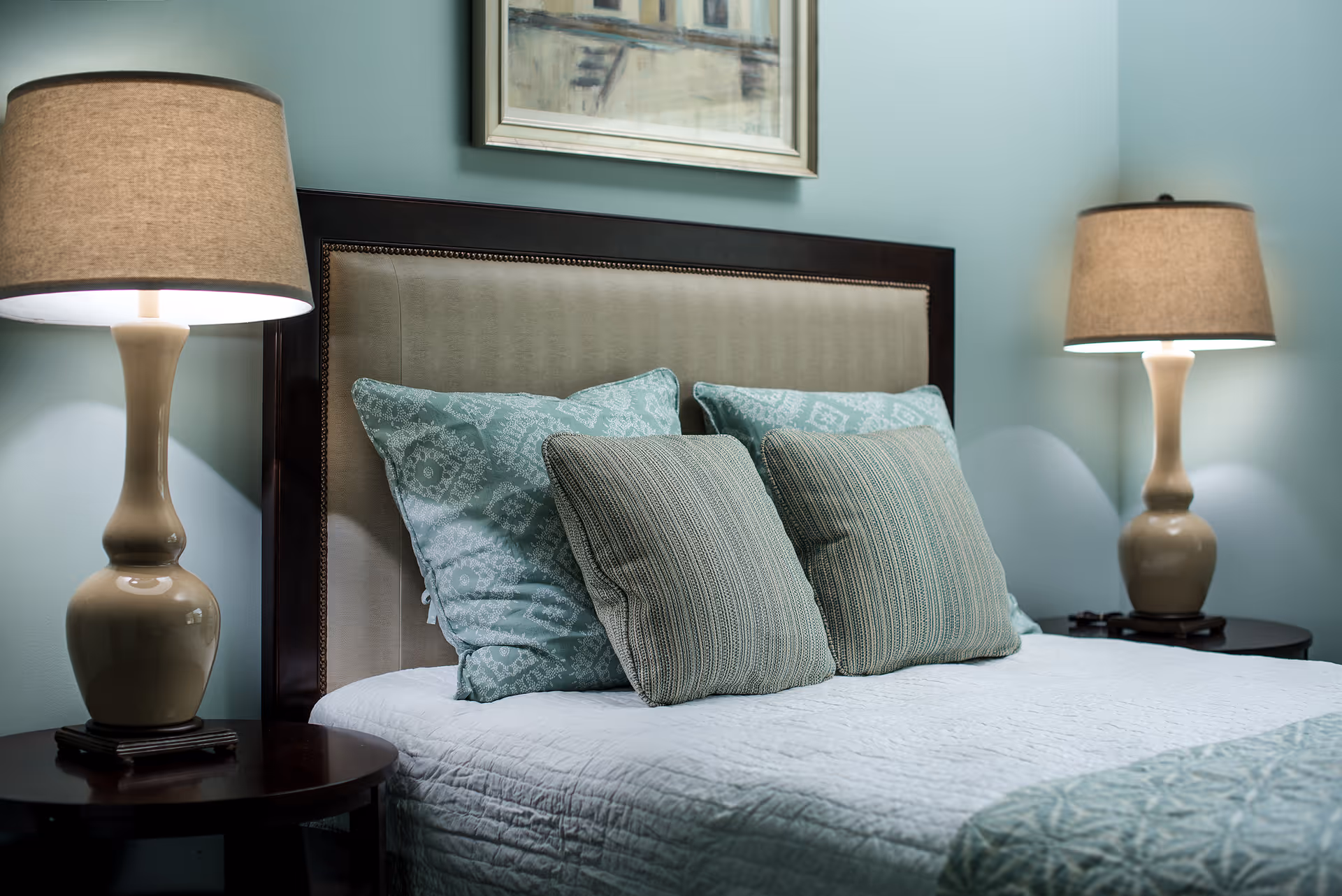 A neatly made bed with decorative green pillows, an upholstered headboard, and matching bedside lamps in a softly lit bedroom.