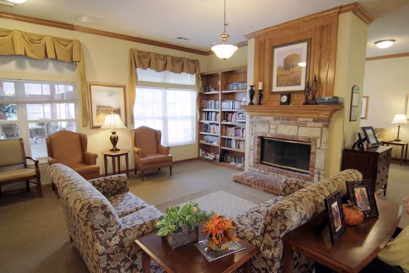 A cozy living room area with floral patterned sofas arranged around a wooden coffee table with plants and flowers. There are two brown armchairs near a window with beige curtains. A brick fireplace with a wooden mantle is on the right side, flanked by built-in bookshelves filled with books. The room is warmly lit with table lamps and a ceiling light fixture.