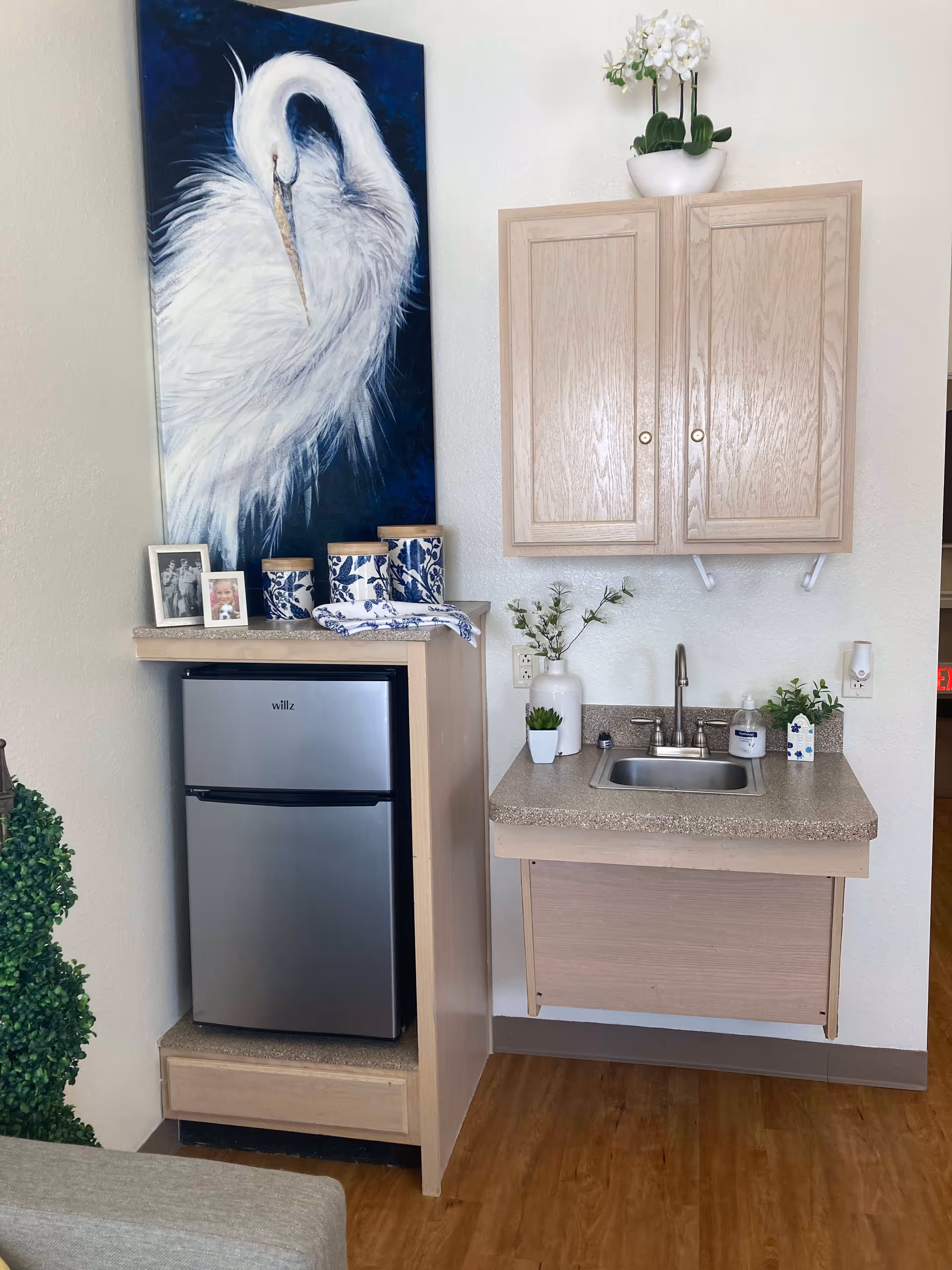 A small kitchenette area featuring a compact stainless steel refrigerator, a countertop with a sink, and light wood cabinets mounted on the wall. Decorative items include a large painting of a white bird, blue and white canisters, framed photos, and small potted plants. The floor is wooden, and the walls are light-colored.