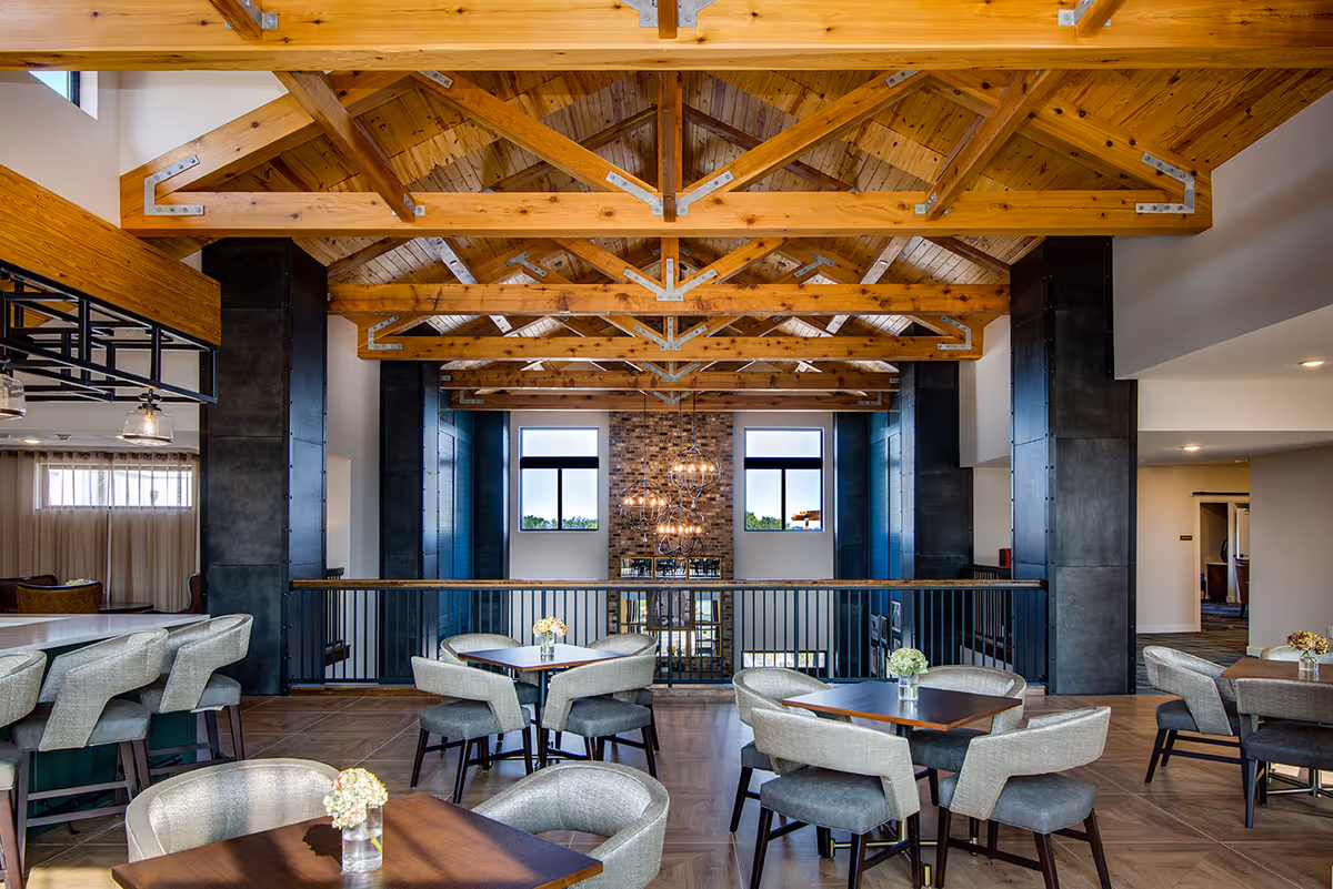 Spacious dining/lounge area with exposed wooden trusses, multiple tables and upholstered chairs, and large windows with a central chandelier.