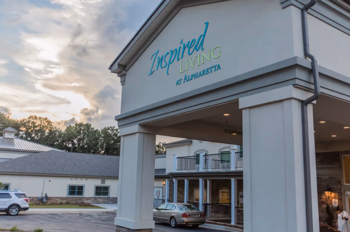 Entrance canopy and facade of the Inspired Living at Alpharetta senior living building with parked cars and a cloudy sky.