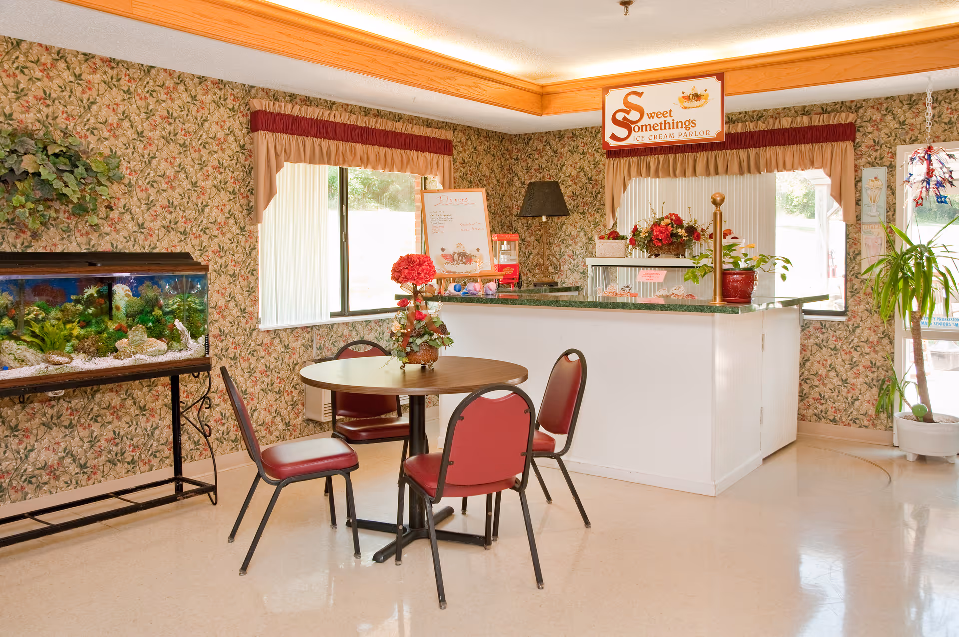 A small ice cream parlor area inside a facility with floral wallpaper, a round table with four red chairs, a fish tank on a stand, and a counter with a sign that reads 'Sweet Somethings Ice Cream Parlor'. The counter has flowers and a menu board, with windows letting in natural light.