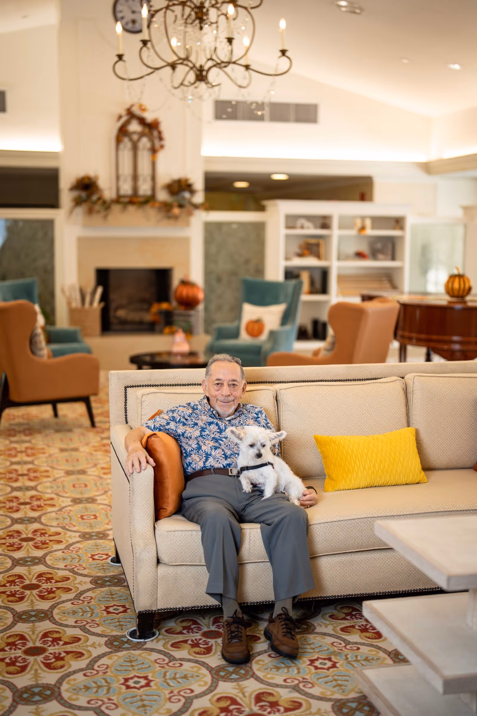 An elderly man sitting on a beige couch in a cozy living room, holding a small white dog on his lap. The room features a patterned carpet, a chandelier, a fireplace decorated with autumn-themed items, and comfortable armchairs in the background.