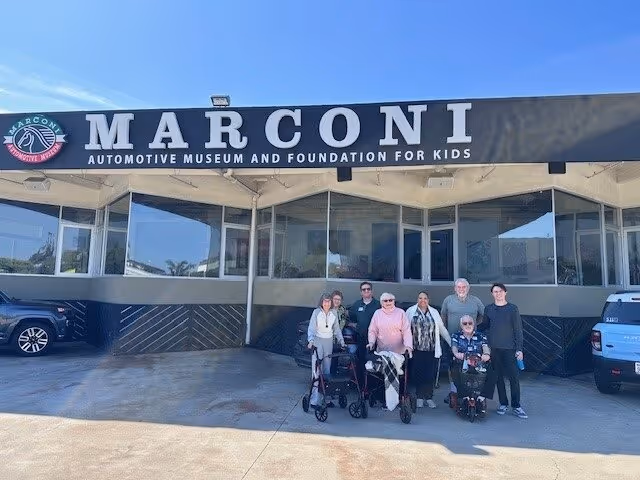 Seven people, including seniors using walkers and a mobility scooter, stand in front of the Marconi Automotive Museum building.