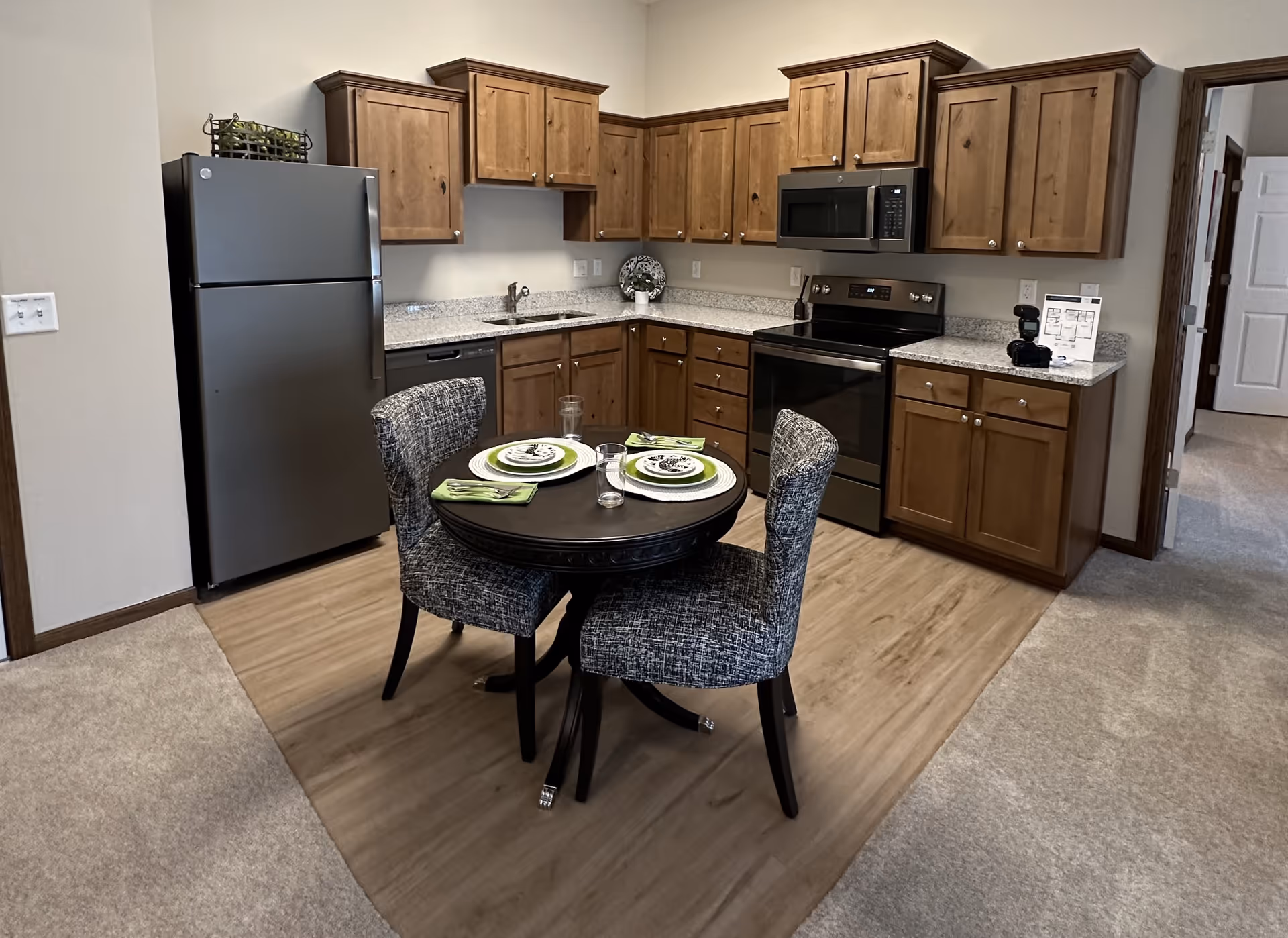 A kitchen area with wooden cabinets, a stainless steel refrigerator, stove, microwave, and dishwasher. In front of the kitchen is a small round dining table set with two place settings and two upholstered chairs. The floor transitions from carpet to wood-like flooring in the kitchen area.