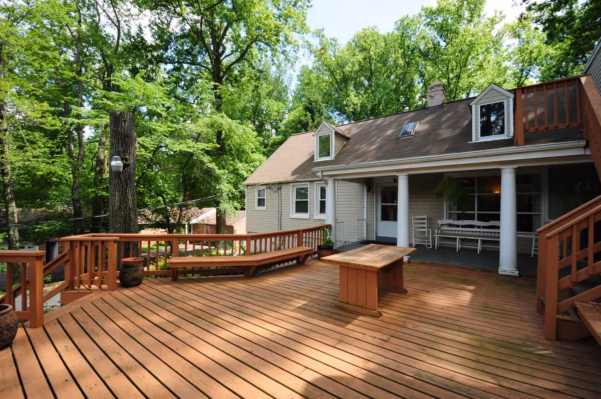 A spacious wooden deck attached to a house with beige siding and a brown roof. The deck features built-in wooden benches, a wooden table, and potted plants. The house has white columns supporting a covered porch area with white rocking chairs and a bench. The surrounding area is lush with green trees and foliage.