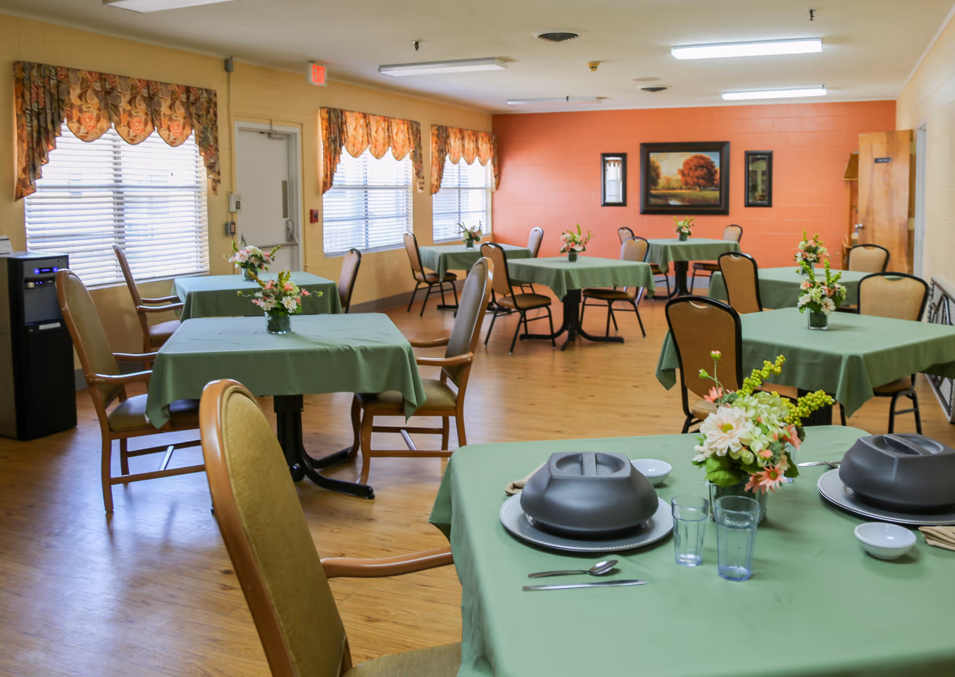 A bright dining room with multiple tables covered in green tablecloths, chairs, and floral centerpieces.