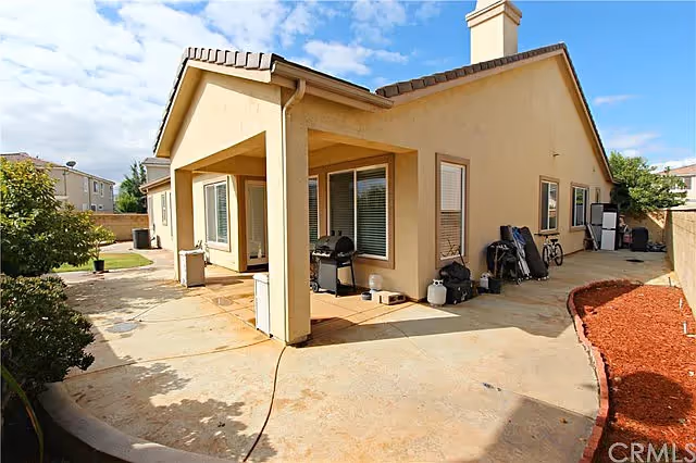 Outdoor view of a single-story beige stucco house with a covered patio area. The patio has a barbecue grill and various items stored against the walls. The yard features a concrete surface with some landscaping including a mulched area and bushes. The sky is partly cloudy.