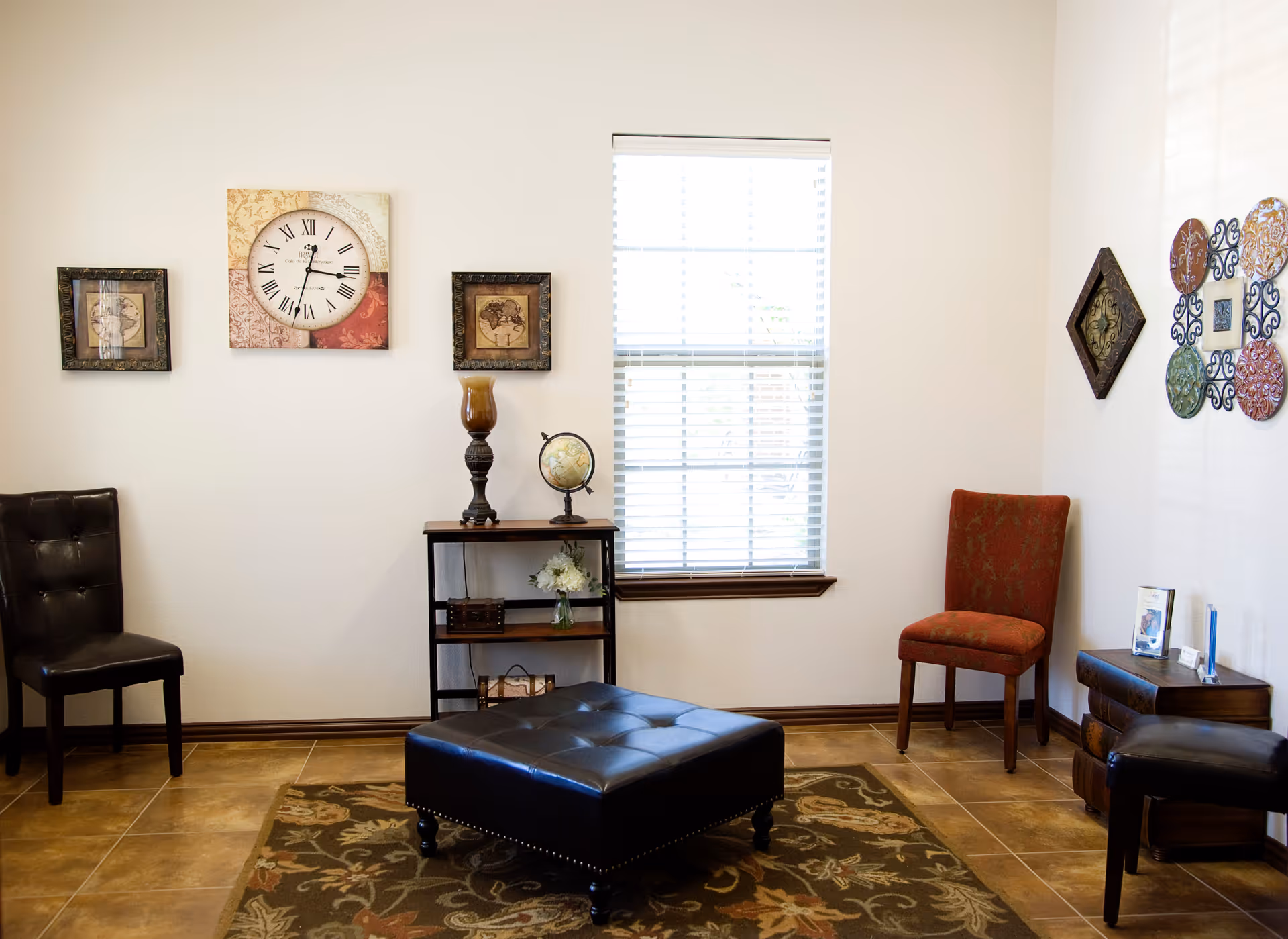 A cozy living room area with a large window with blinds, two chairs (one black and one red), a black leather ottoman on a patterned rug, a small wooden shelf with decorative items including a globe and a lamp, and various framed wall decorations including a clock and colorful metal art pieces.