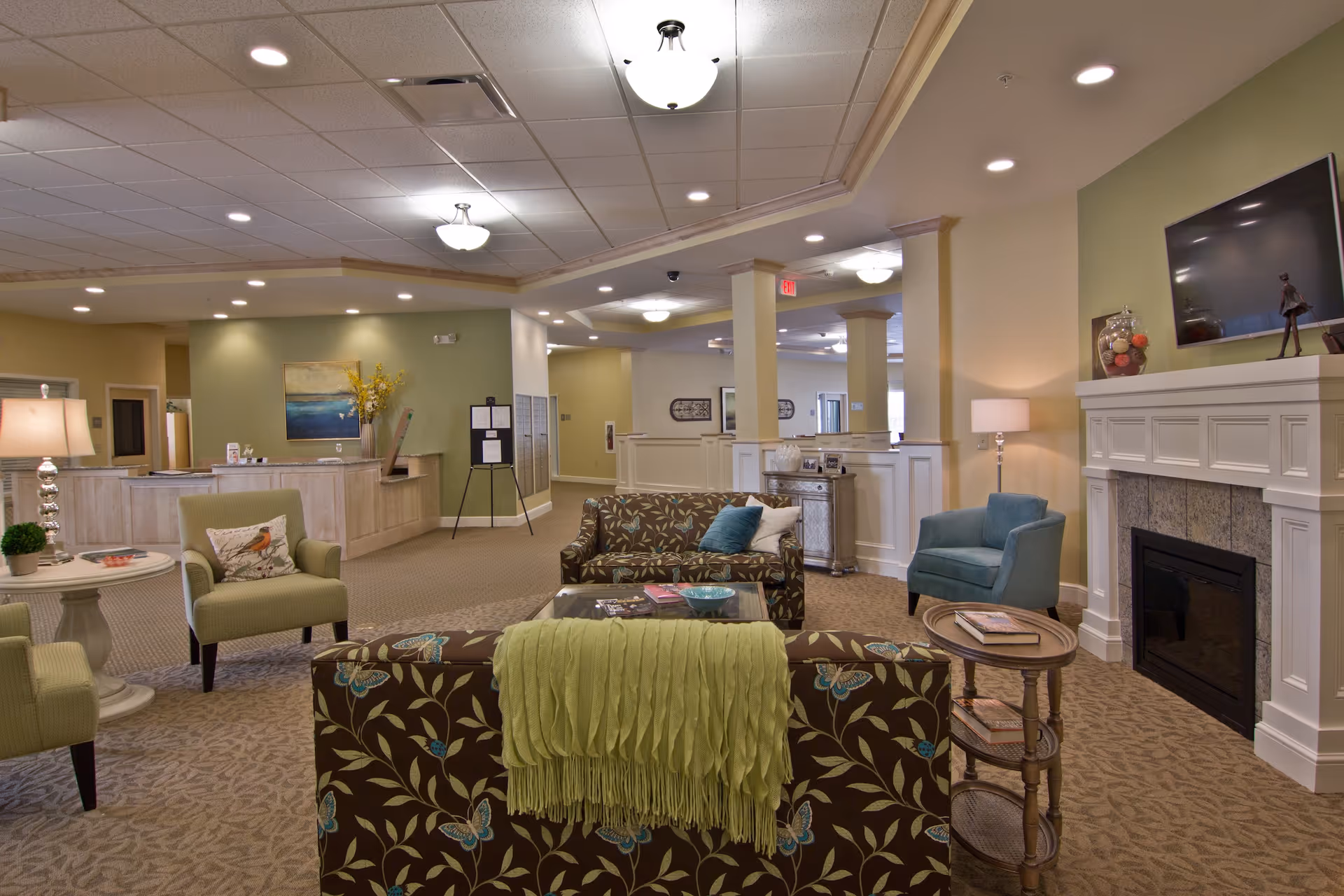 A cozy senior living facility common area with patterned sofas, armchairs, a glass coffee table, a fireplace with a mounted TV above it, and soft lighting. The space has beige carpet, light green and cream walls, and decorative elements including a lamp, artwork, and a small side table with books.