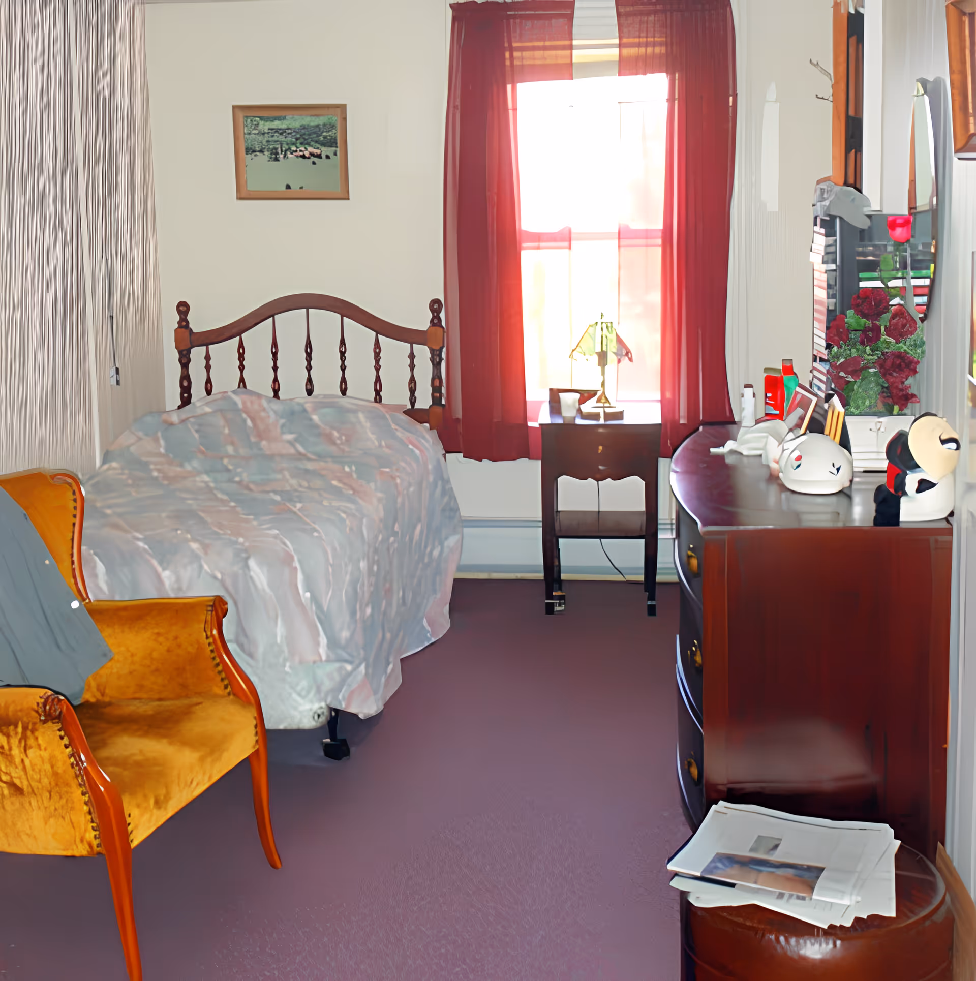 A small bedroom with a single bed covered in a patterned blanket, a wooden headboard, a yellow upholstered armchair, a wooden dresser with various items on top including a lamp and decorative objects, a small side table with a lamp in front of a window with red curtains, and a framed picture on the wall.