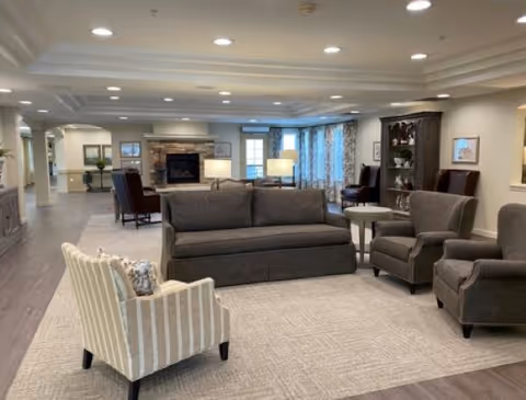 A spacious and well-lit senior living community lounge featuring a gray sofa, two matching gray armchairs, and a striped armchair arranged around a light-colored area rug. In the background, there is a stone fireplace with a mantel, additional seating, lamps, and large windows with curtains allowing natural light to fill the room.