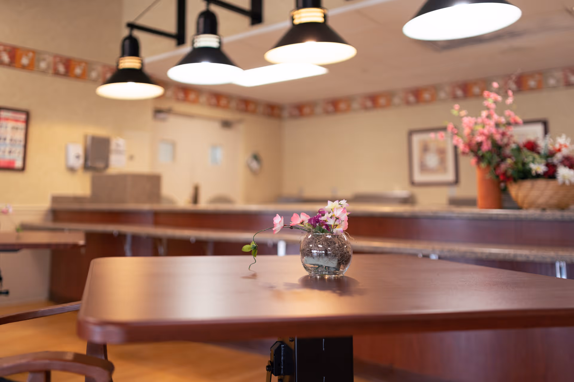 A dining area with a wooden table in the foreground holding a small glass vase with pink and white flowers. In the background, there are more tables, a counter with additional flower arrangements, framed pictures on the wall, and hanging ceiling lights.