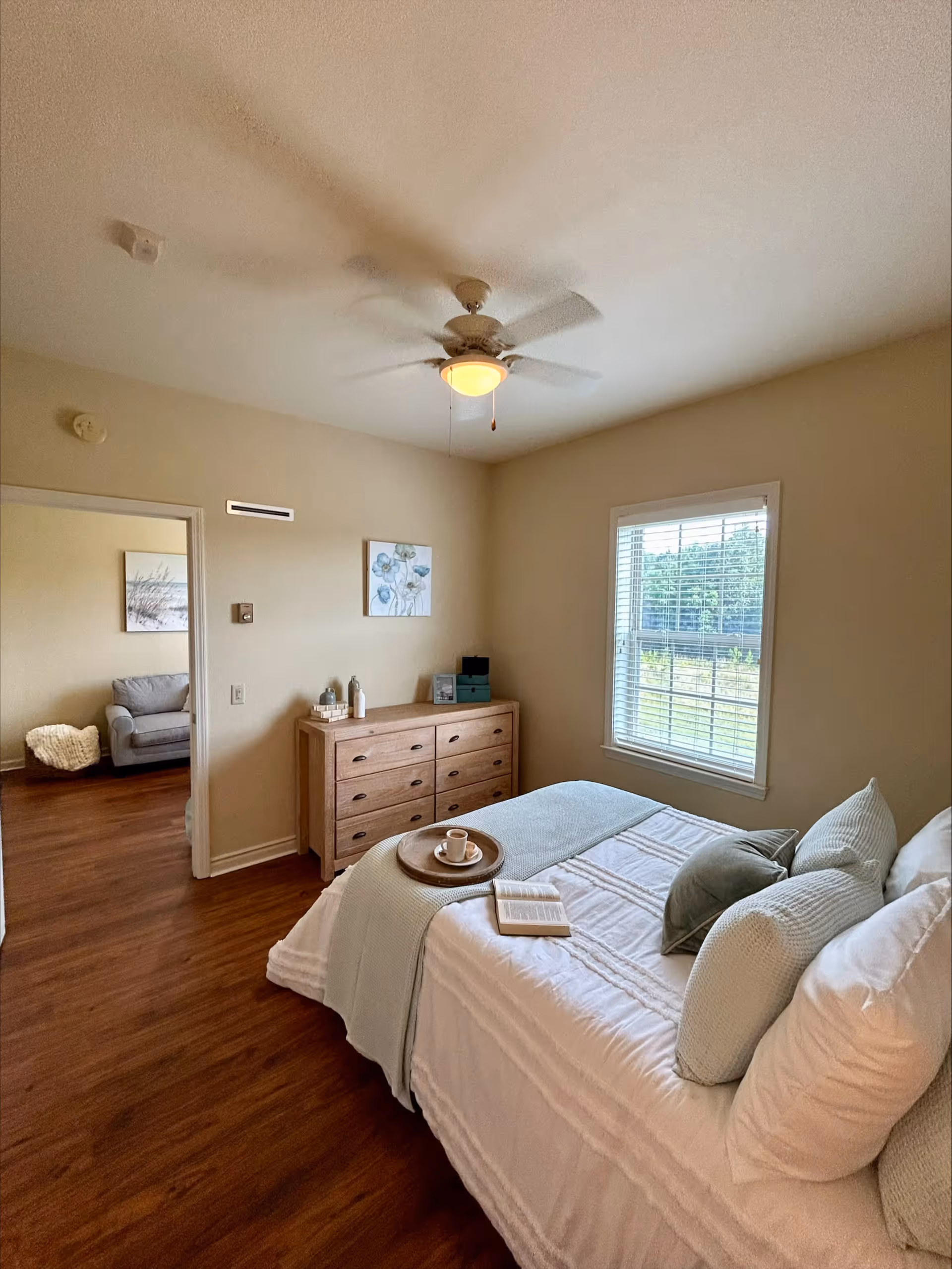 A cozy bedroom with a neatly made bed featuring white and light green bedding and pillows. A wooden dresser with decorative items sits against the wall, and a window with blinds lets in natural light. A ceiling fan with a light fixture is on the ceiling. Through an open doorway, a glimpse of a living area with a gray armchair and a basket is visible.