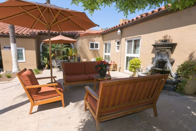 Outdoor patio area with cushioned wooden seating including a sofa and two chairs, two large umbrellas providing shade, potted plants, and a decorative wall fountain attached to a beige building under a clear blue sky.
