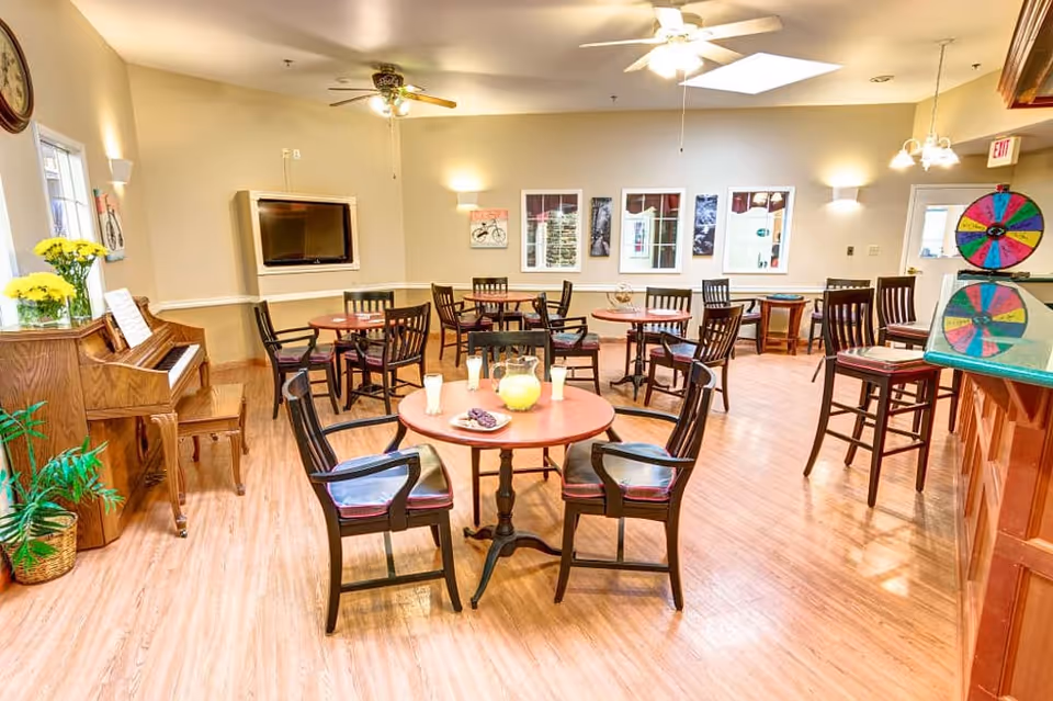 A bright and spacious dining area with several round wooden tables and chairs arranged neatly. A wooden piano with a bench and a vase of yellow flowers is placed against the left wall. The room has light-colored walls, wooden flooring, ceiling fans, and multiple light fixtures. A flat-screen TV is mounted on the far wall, and a colorful spinning wheel is visible on the right side near a counter with bar stools.