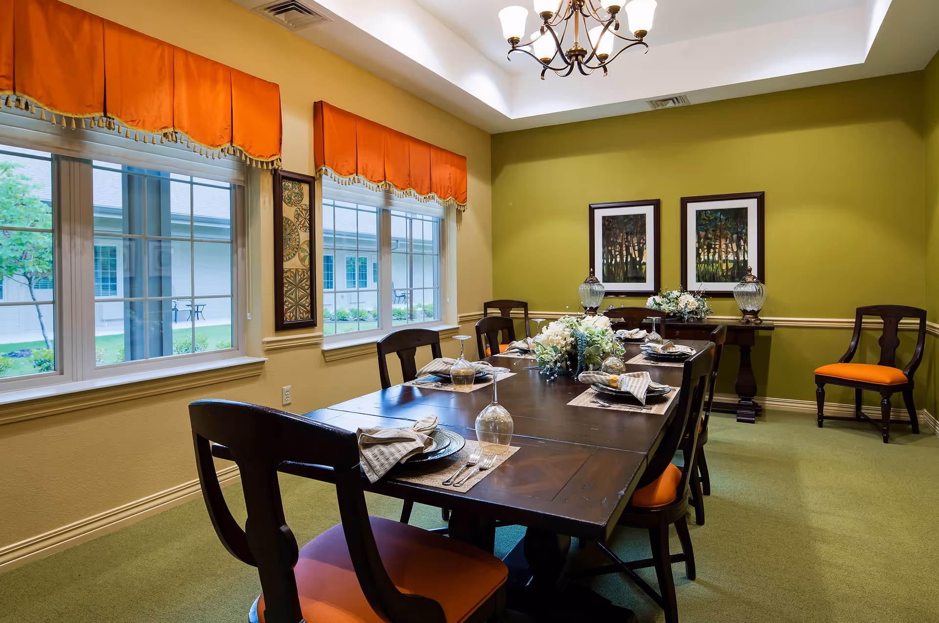 Dining room with a long dark wood table set for a meal, orange-cushioned chairs, green walls, and large windows with orange valances.