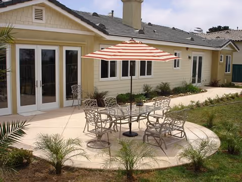 Outdoor patio with a round concrete seating area, metal table and chairs under a striped umbrella in front of a single-story building.