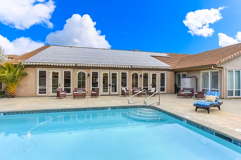 Outdoor swimming pool area with clear blue water in front of a single-story building with multiple glass doors and windows. The building has a tiled roof with solar panels. There are several lounge chairs and seating arrangements around the pool under a bright blue sky with some clouds.