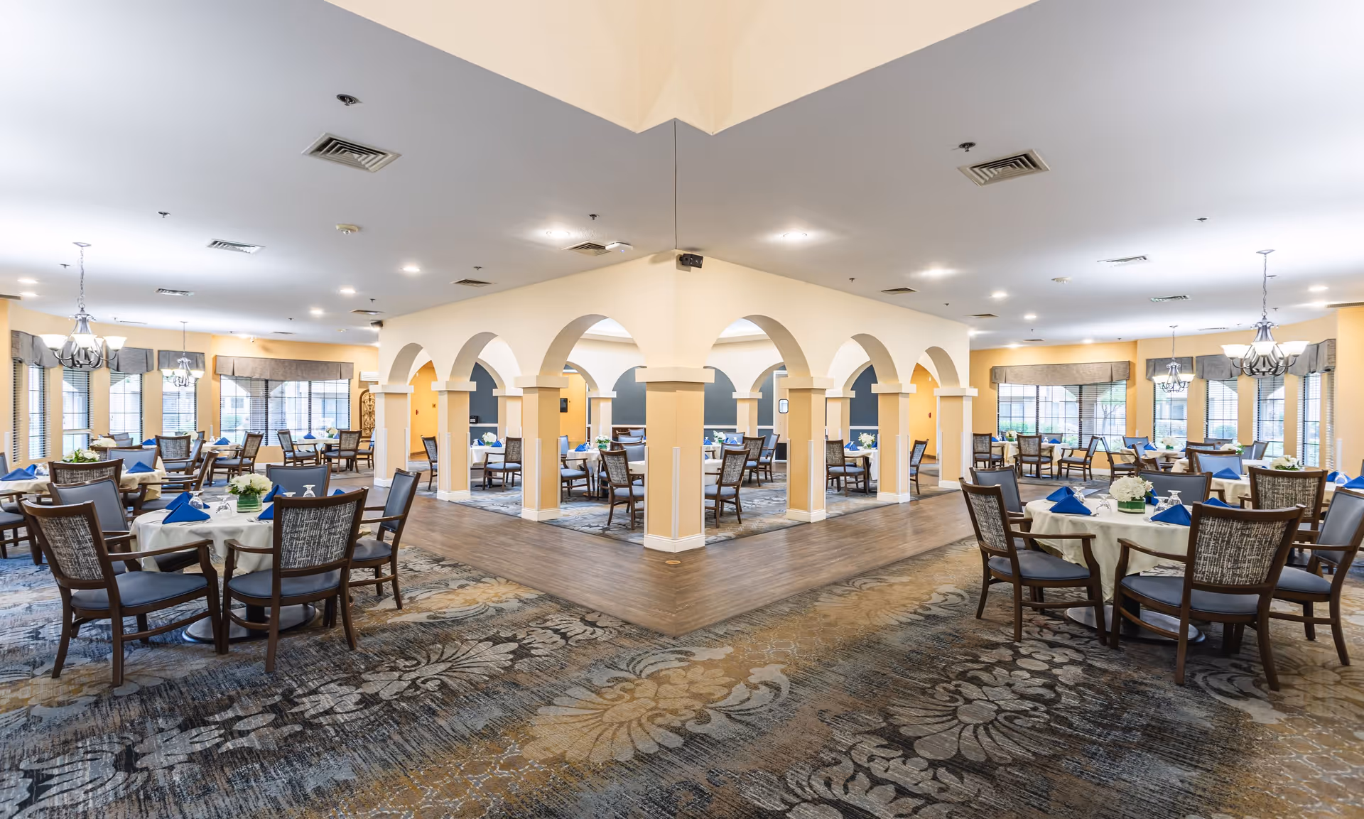 Spacious dining room with multiple round tables covered with white tablecloths, each set with blue napkins and floral centerpieces. The room features large windows with valances, chandeliers, and a decorative carpet with floral patterns. The area is divided by a central structure with arches and columns.