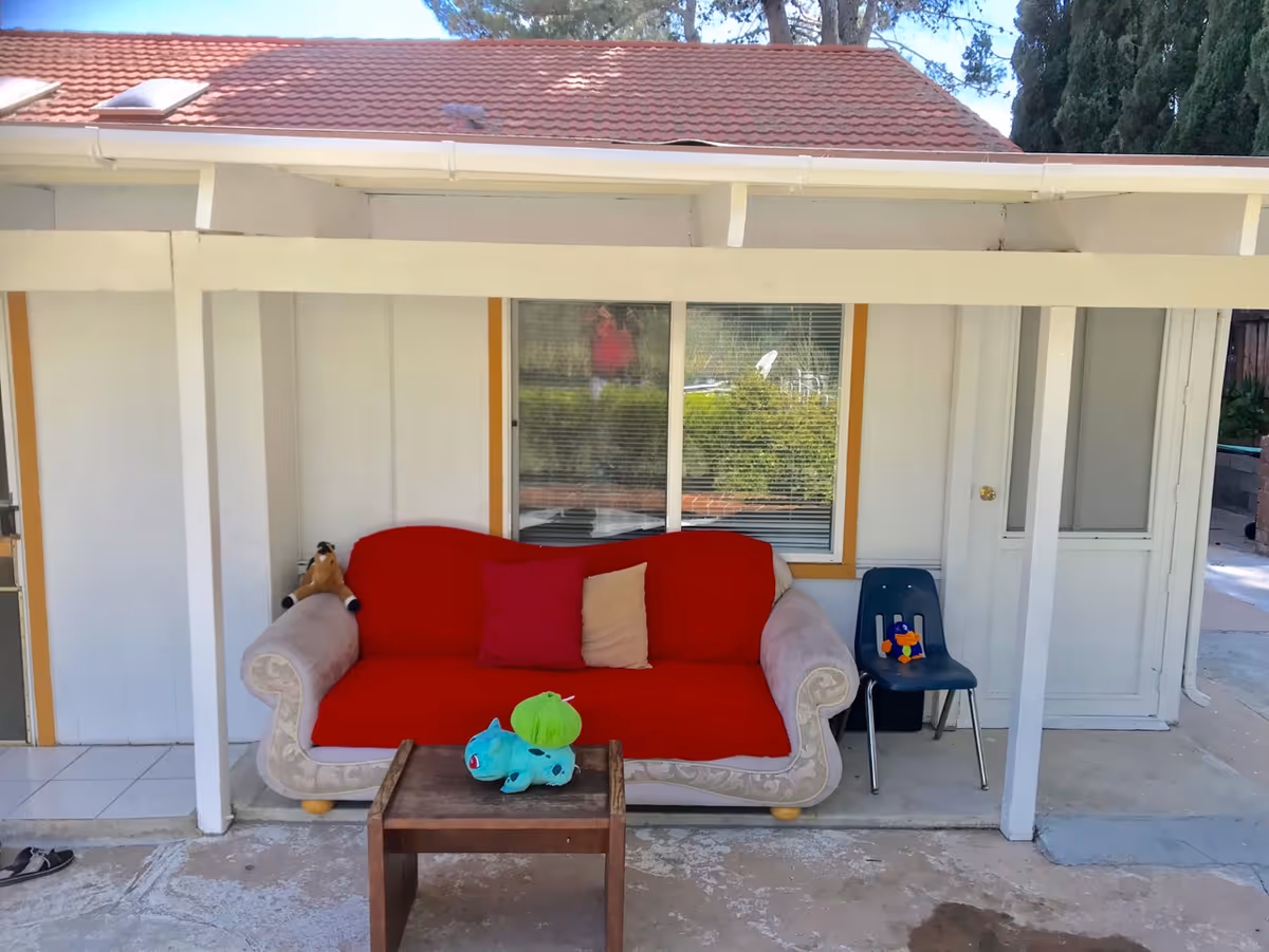 Covered front porch with a red-covered sofa, small wooden table, and a plastic chair in front of a single-story building.