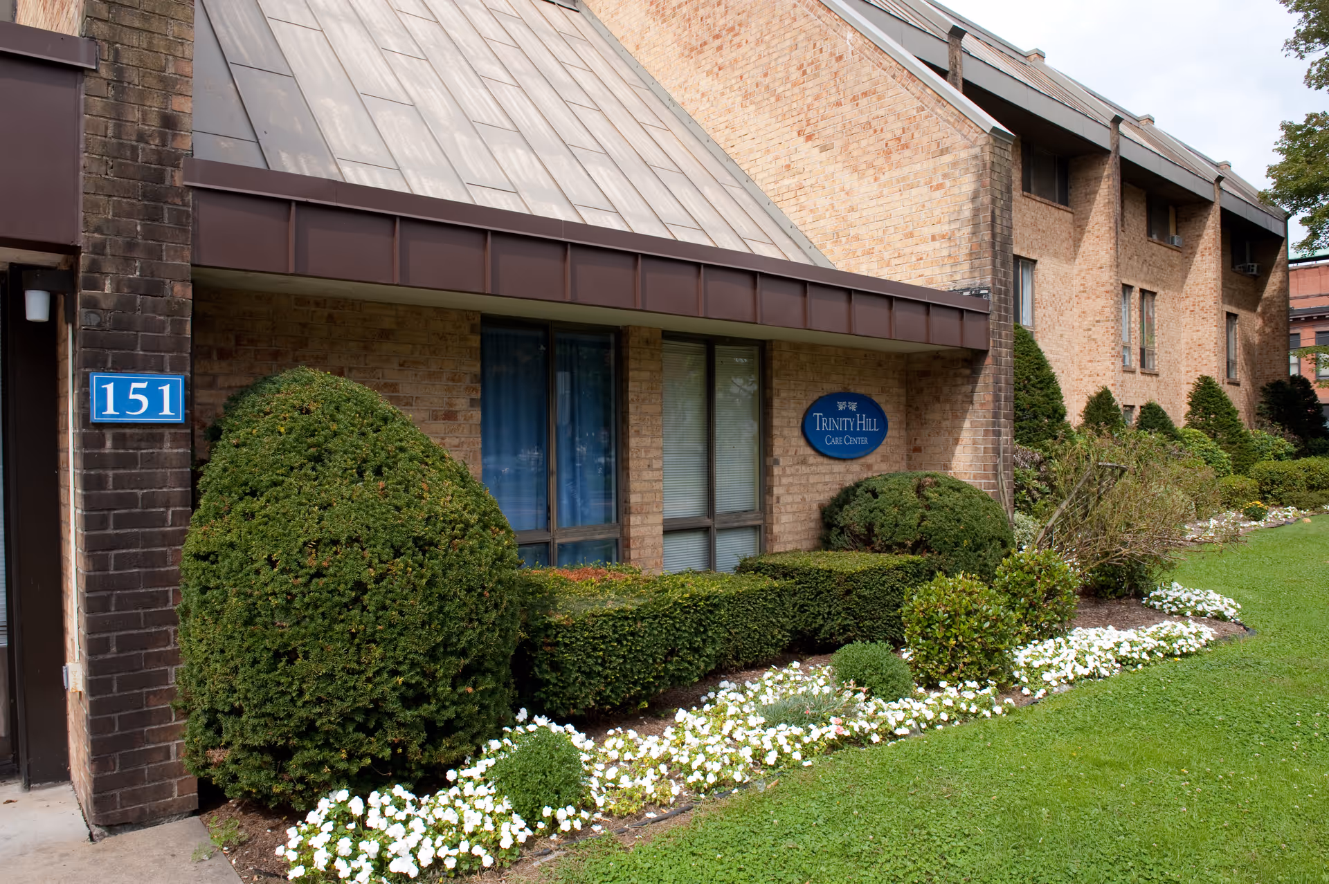 Front exterior of a brick care facility with trimmed shrubs, flowerbeds, a blue "Trinity Hill Care Center" sign, and an address plaque reading "151".