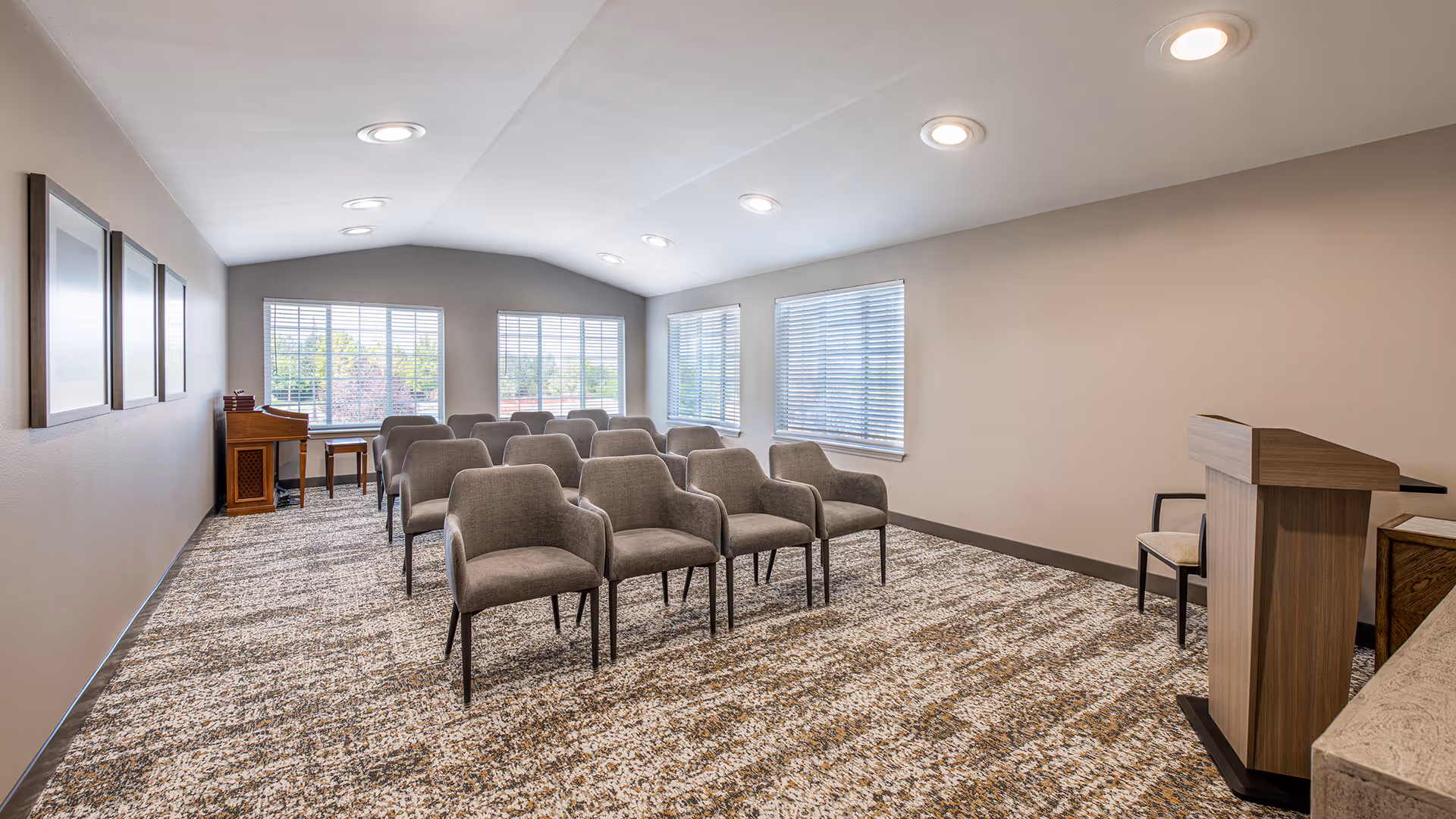 A small conference or meeting room with rows of gray upholstered chairs facing a wooden podium. The room has beige walls, a patterned carpet, and several windows with blinds allowing natural light to enter. There are framed pictures on one wall and a small wooden desk with a chair near the windows.