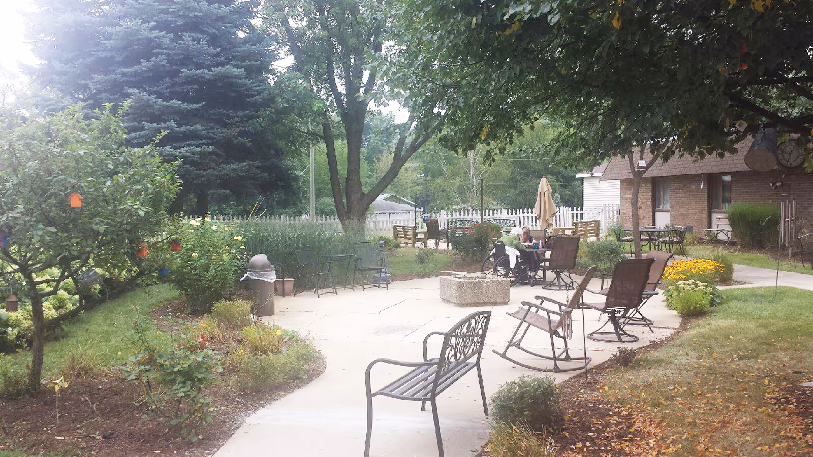 Outdoor patio area at a senior living facility with several chairs and tables arranged on a concrete surface surrounded by grass, trees, and flower beds. A person in a wheelchair is seated at one of the tables. The background includes a white picket fence and a brick building.
