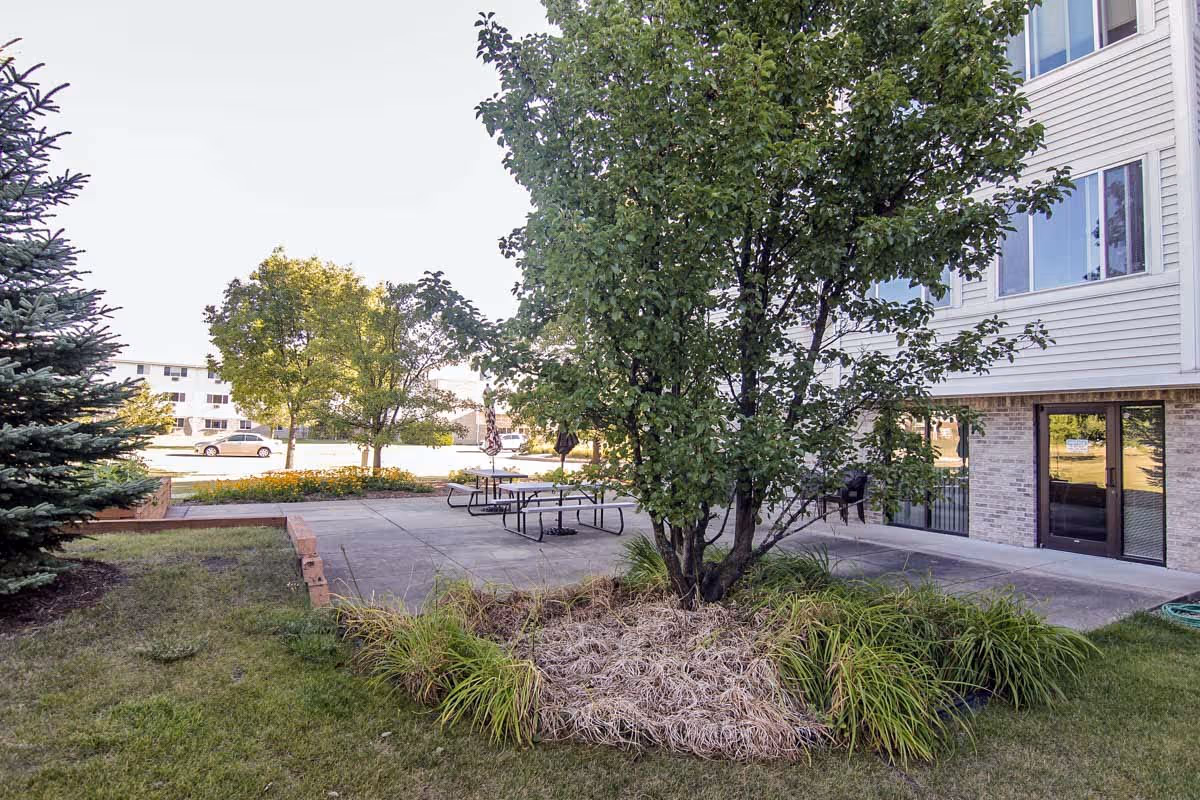 Outdoor patio area at Villa Ciera Senior Apartments featuring a concrete seating area with picnic tables, surrounded by green grass, trees, and landscaping. The side of a multi-story building with windows and a door is visible on the right.
