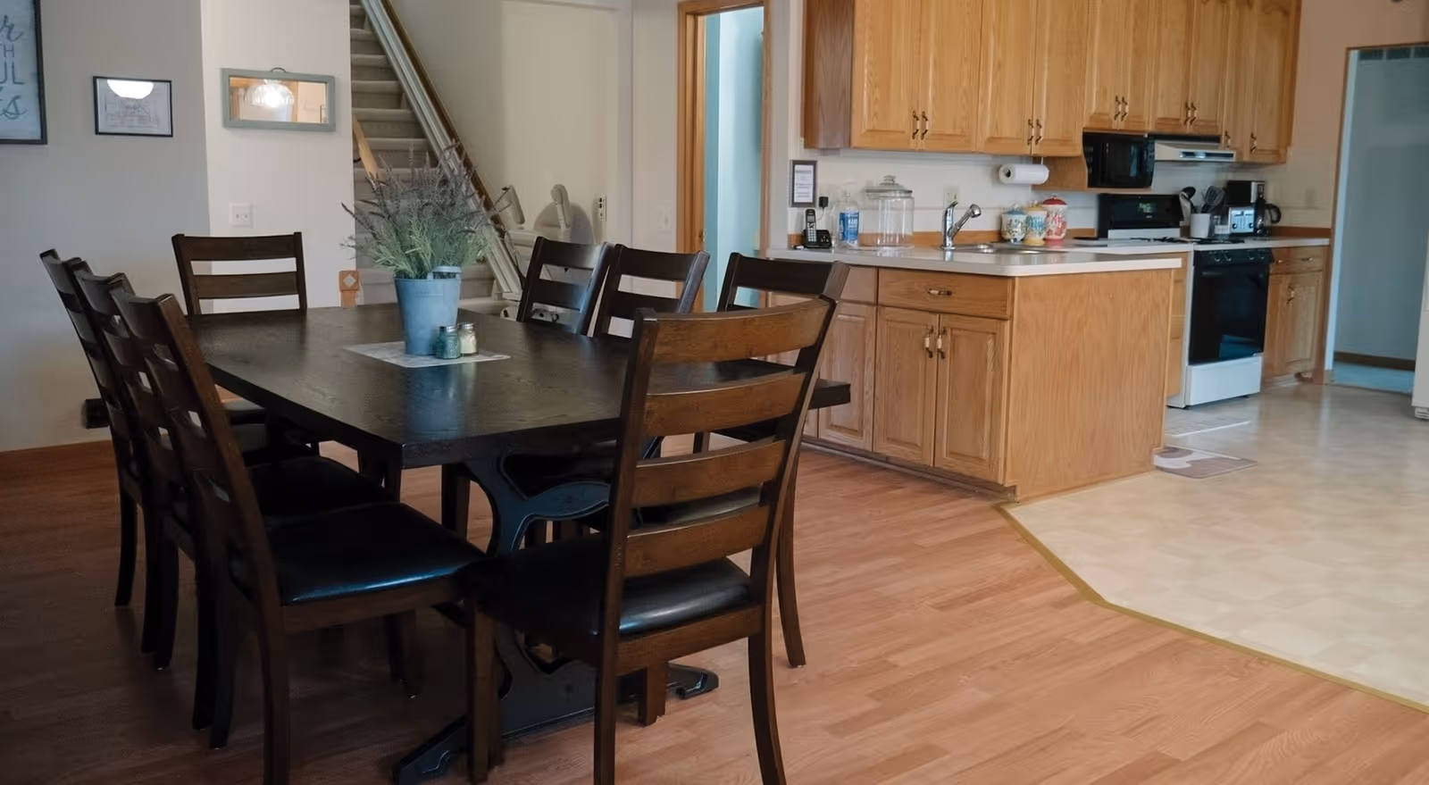 A dining area with a dark wooden table surrounded by eight matching chairs with black cushions. A small plant and salt and pepper shakers are placed on a mat in the center of the table. In the background, there is a kitchen with wooden cabinets, a white stove, a microwave, a coffee maker, and a sink. The floor transitions from wood in the dining area to a light-colored tile in the kitchen. A staircase is visible in the background along with some wall decorations.